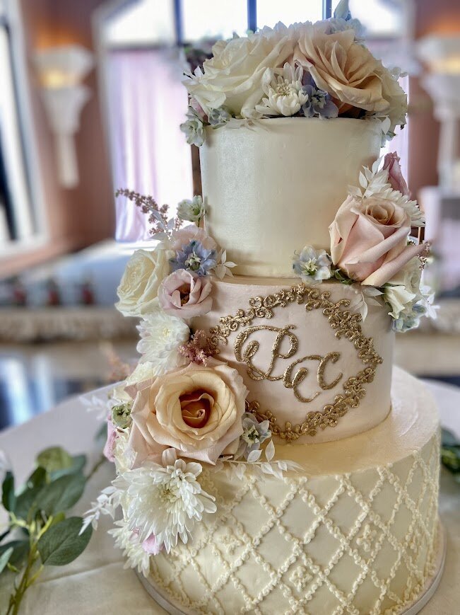 A close up of a tiered cake. The top and bottom tiers are ivory colored frosting and the middle is a pale pink. The bottom tier has a lacey crisscross piped pattern. The middle tier has an ornate monogram in gold with gold filigree in a diamond around it. There's fresh flowers down the left side and filling the top in white and pinks.