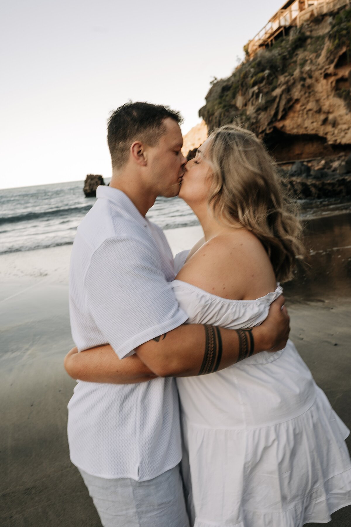 engaged Couple wearing white kiss at taylors mistake in christchurch with the water behind them