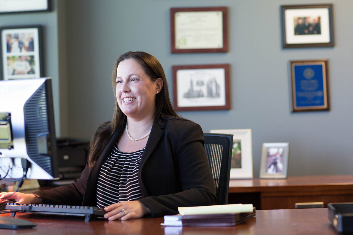 female businesswoman sitting at a wooden desk with diplomas behind her