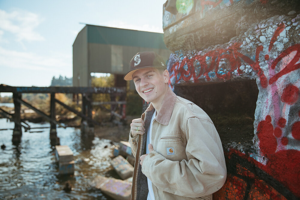 High school boy smiling for his senior pictures with a large jacket and baseball cap
