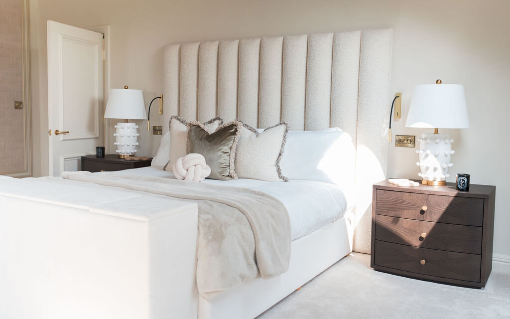 A well-lit bedroom featuring a large plush bed with a tall, tufted headboard. The bed is flanked by two dark wooden nightstands, each with a white lamp. The room has a neutral color palette with white and beige bedding and some accent pillows.