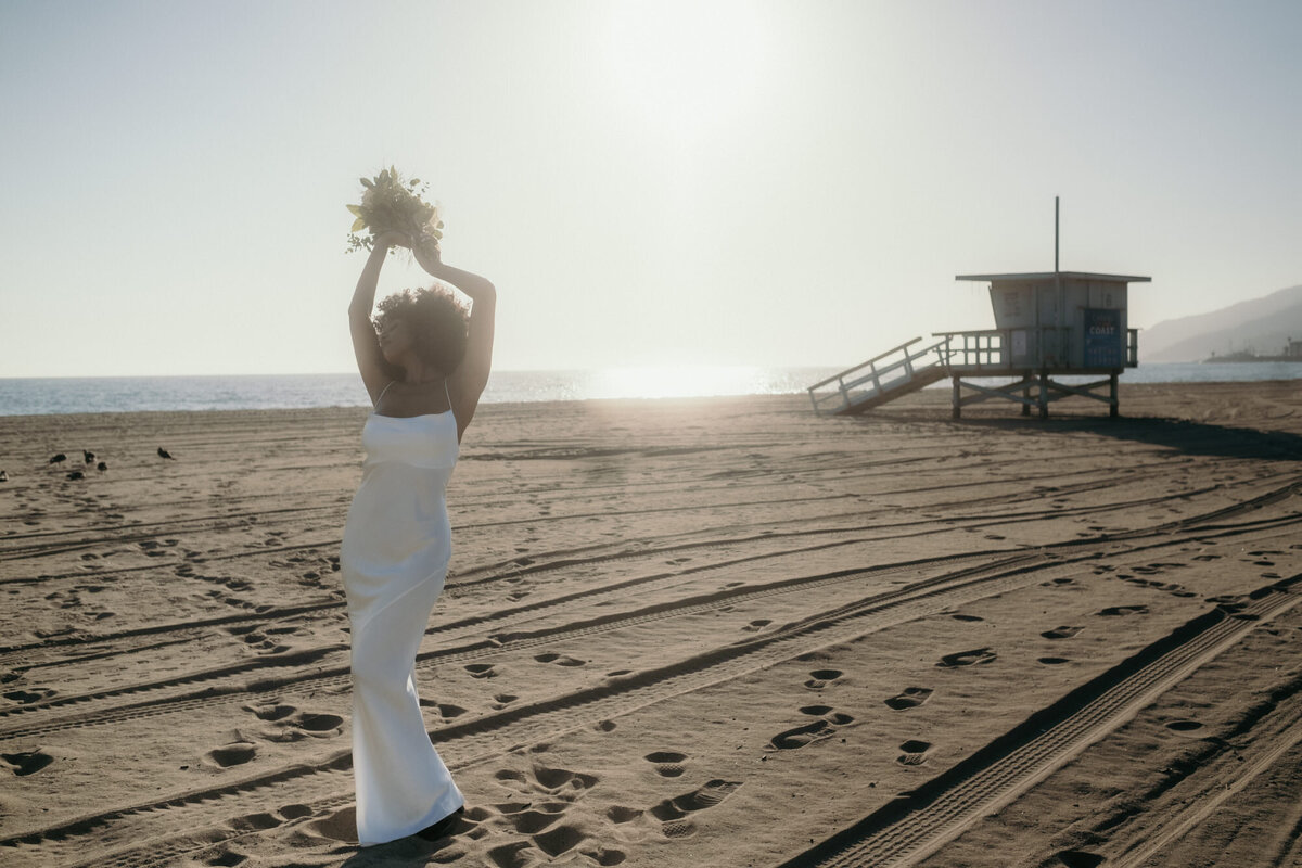 Von der Ferne betrachtet hält die Braut im Sonnenschein am Strand ihren Strauß hoch.