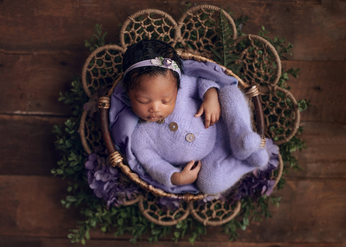 little sleepy newborn wearing soft lilac knit outfit and headband, posed in a rattan bow on a decorative bow with greenery and purple flowers