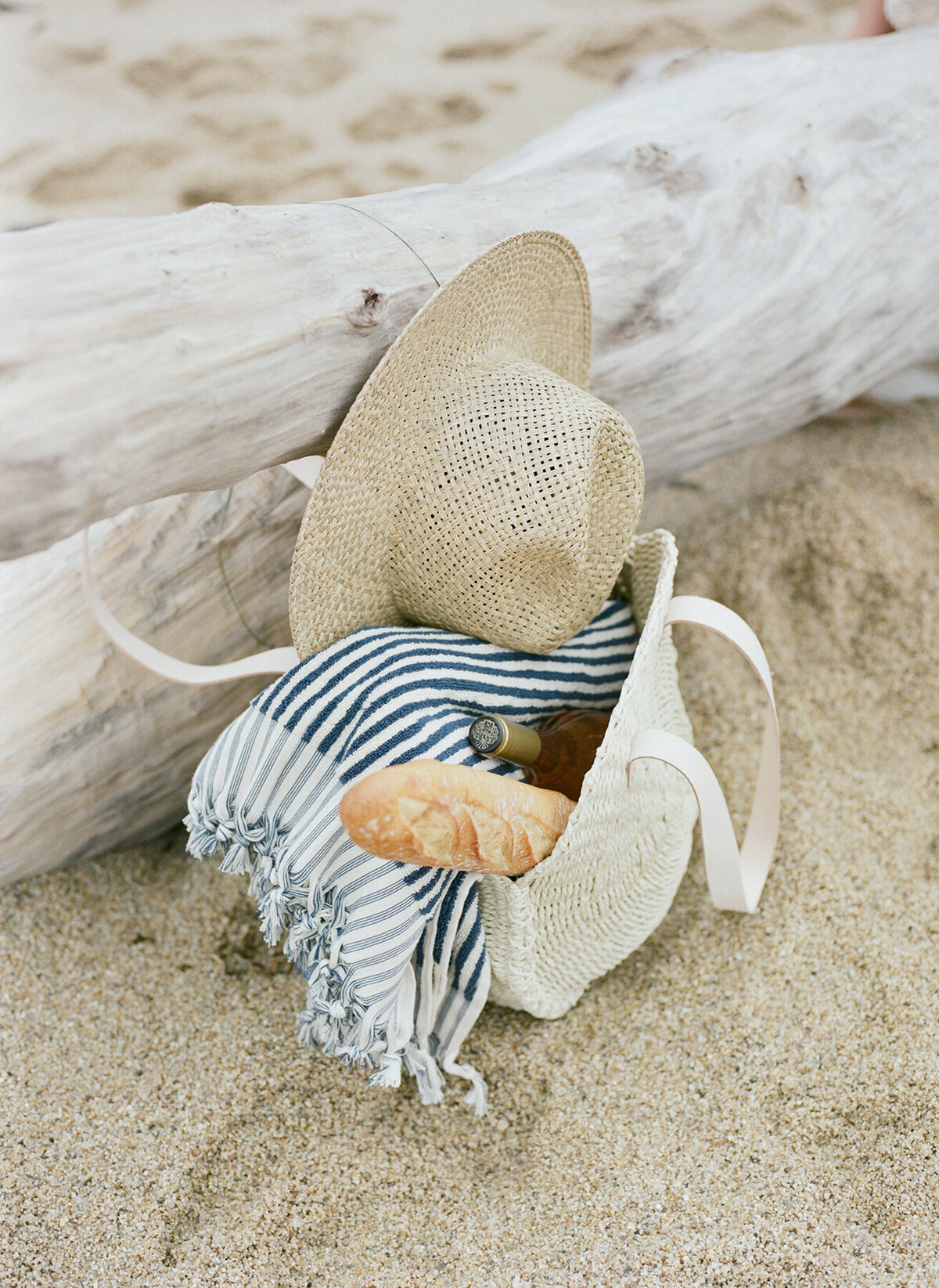 basket of a towel, sun hat, wine, and baguette on the sand by piece of drift wood