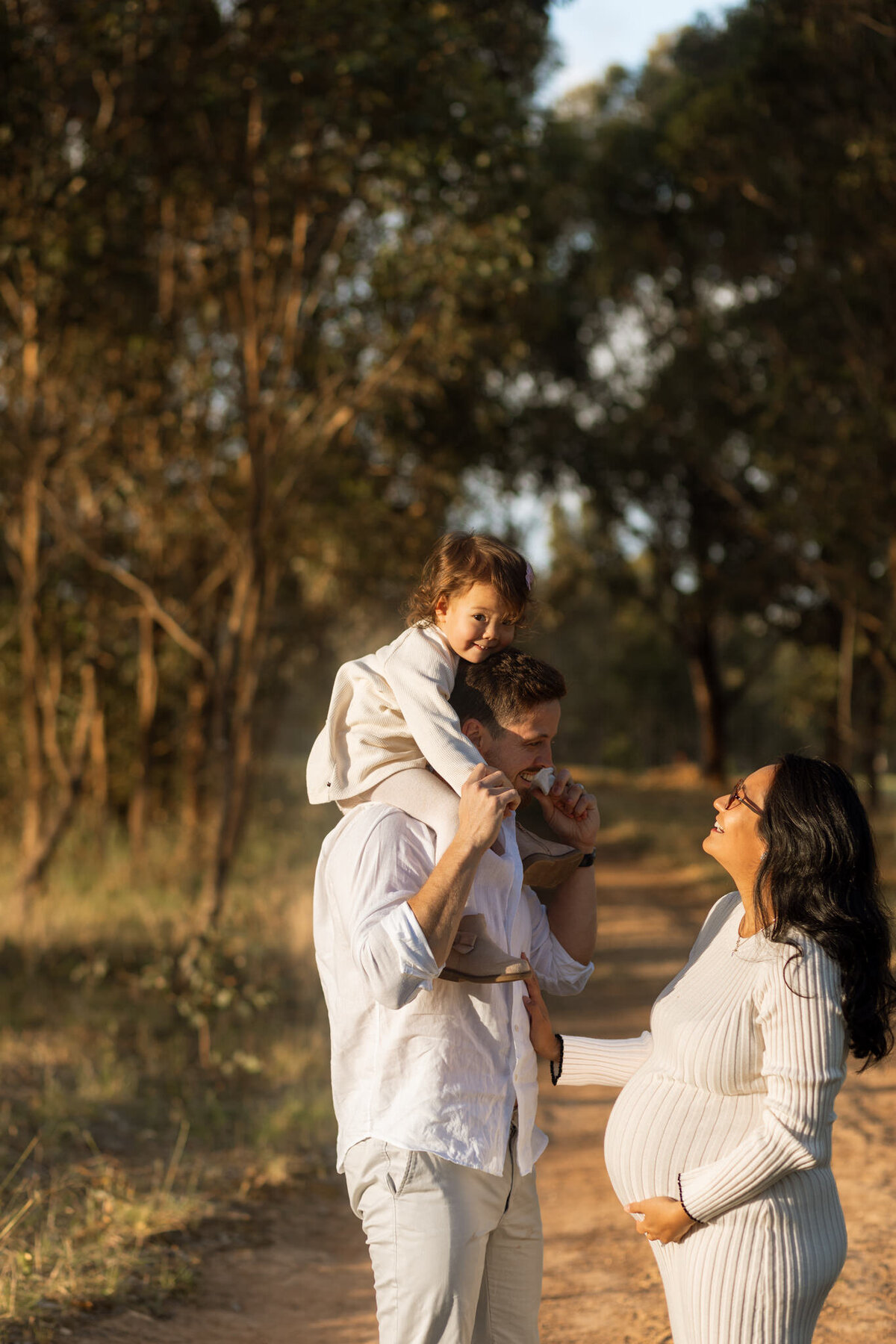 Maternity photoshoot in Sydney featuring a pregnant mother smiling at her partner and daughter during golden hour in a natural outdoor setting