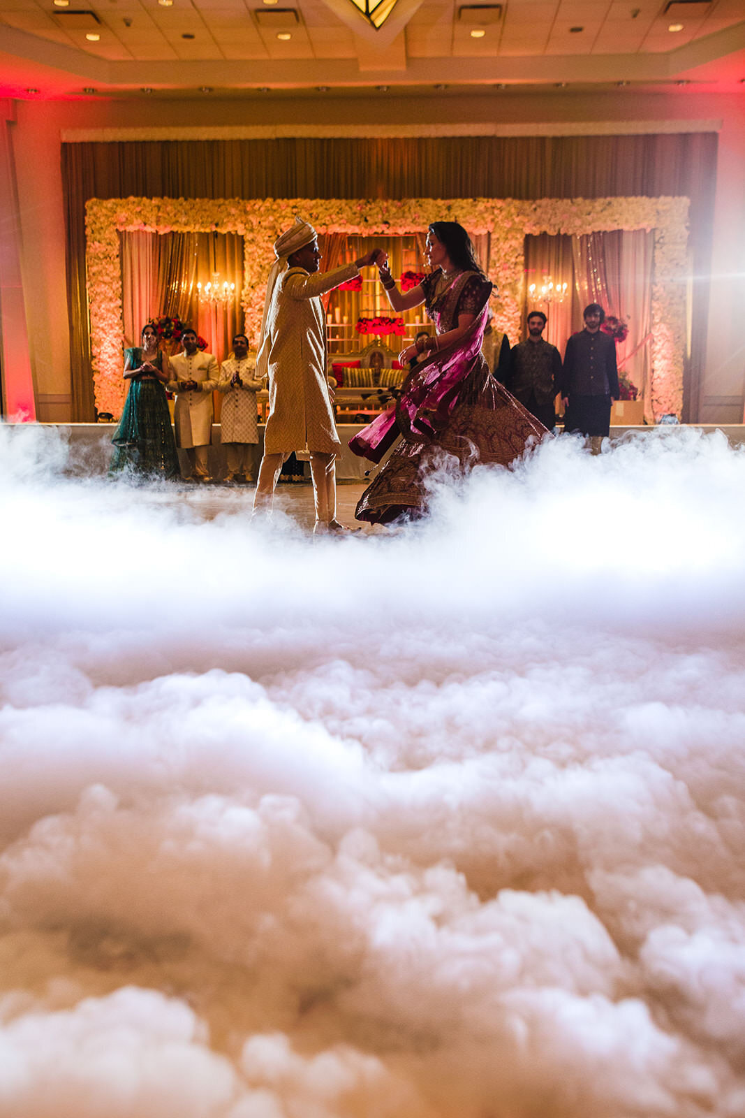A bride in a dark red lehenga and groom in a sherwani are surrounded by a foggy effect on the dance floor