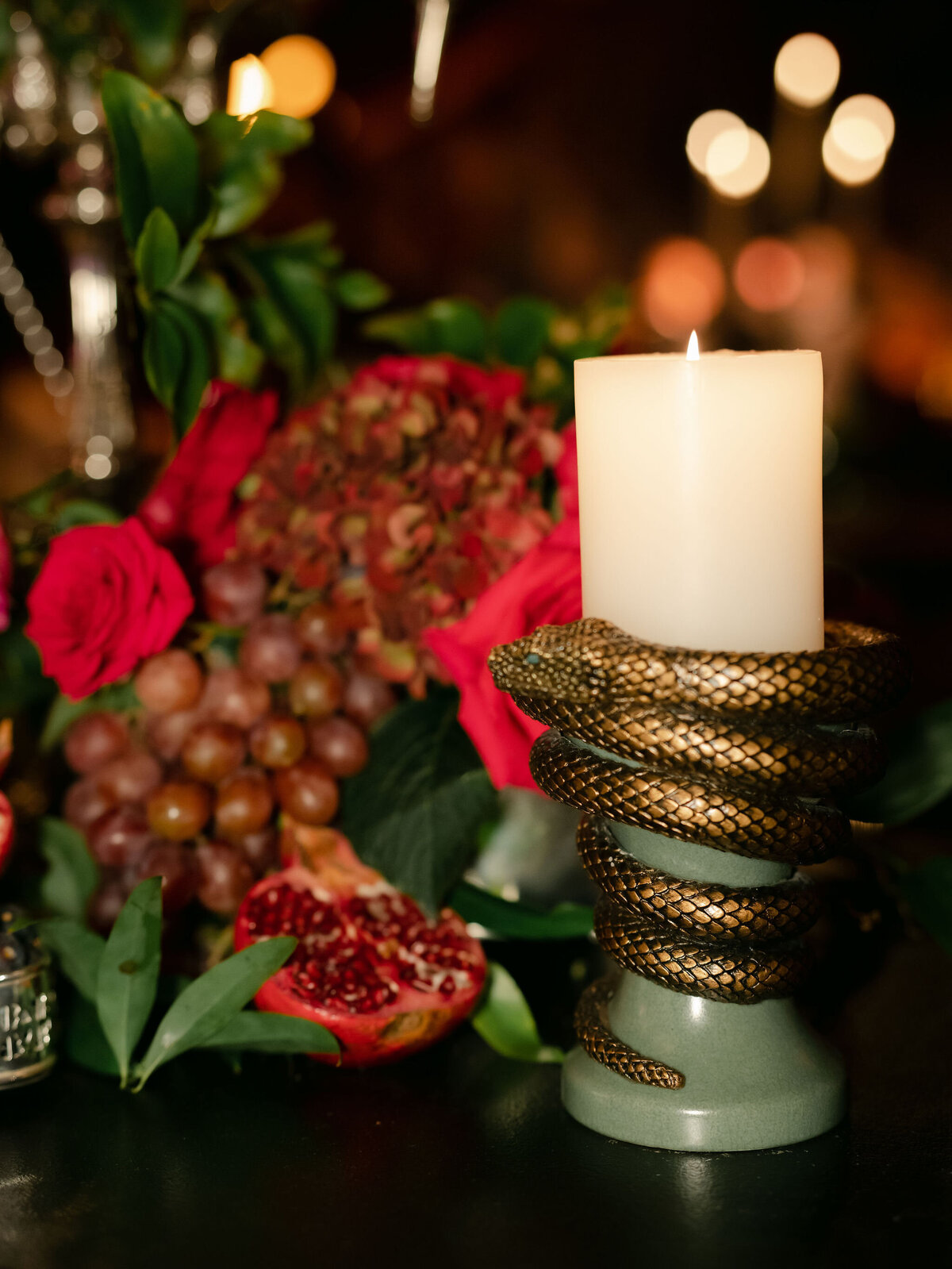 The photo was taken at a beautiful tented wedding in Virginia captured by the talented photographer Elisabetta Marzetti, with videographers Clayton Films also present. In the image, a lit white candle is delicately held by a golden snake-shaped holder, artfully arranged amidst a backdrop of vibrant flowers, grapes, and a split pomegranate. The soft, warm lighting in the scene enhances its cozy and inviting ambiance, making it perfect for a festive or romantic setting.