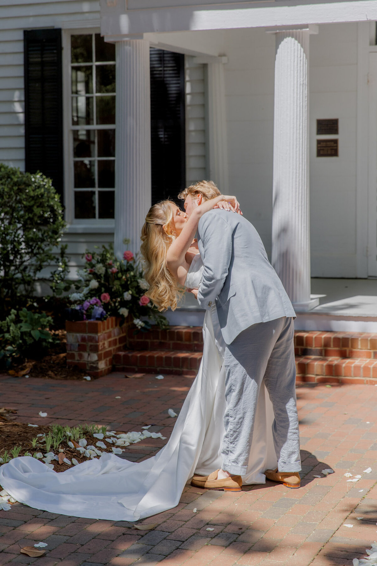A bride and groom kiss on their wedding day. Behind them is a simple white house.