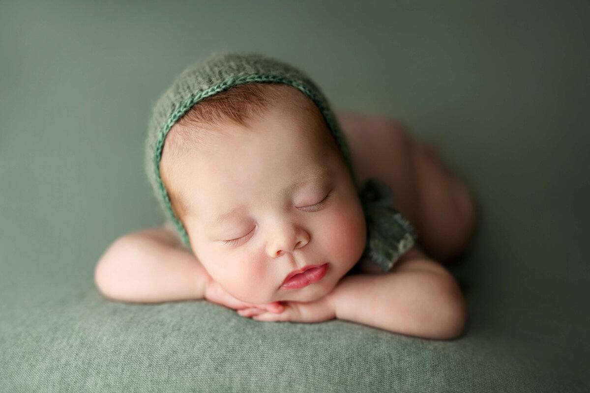 Newborn girl laying on her tummy  on a green backdrop with matching bonnet at a baby photo shoot in Northern VA