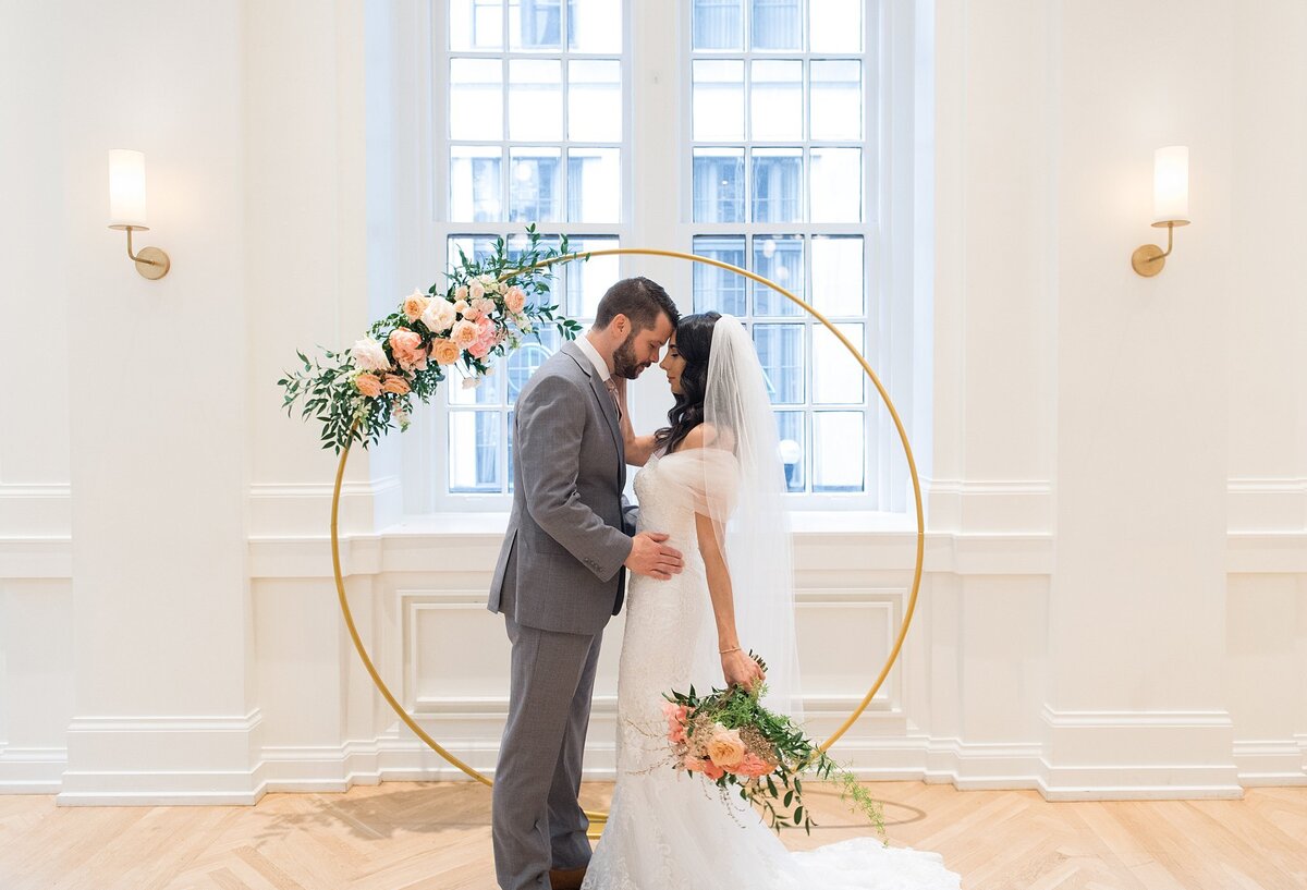 A bride wearing a flowing off the shoulder wedding dress with a long veil kisses the groom wearing a grey suit in front of their gold circular arbor. The bride holds a cascading bouquet of pink peonies, ivory roses and greenery which matches the floral spray of pink, peach and ivory flowers on the arbor. The Noelle Hotel ballroom has soft white walls, tall arched windows and blond hardwood floors.