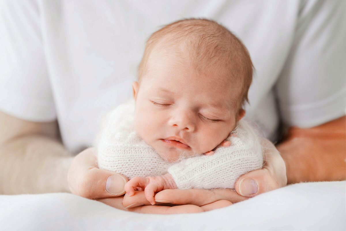 A newborn in a white fuzzy sweater is laying on her tummy in her father's hands in a photo taken by Val Stone Photography in an Apex NC Studio.