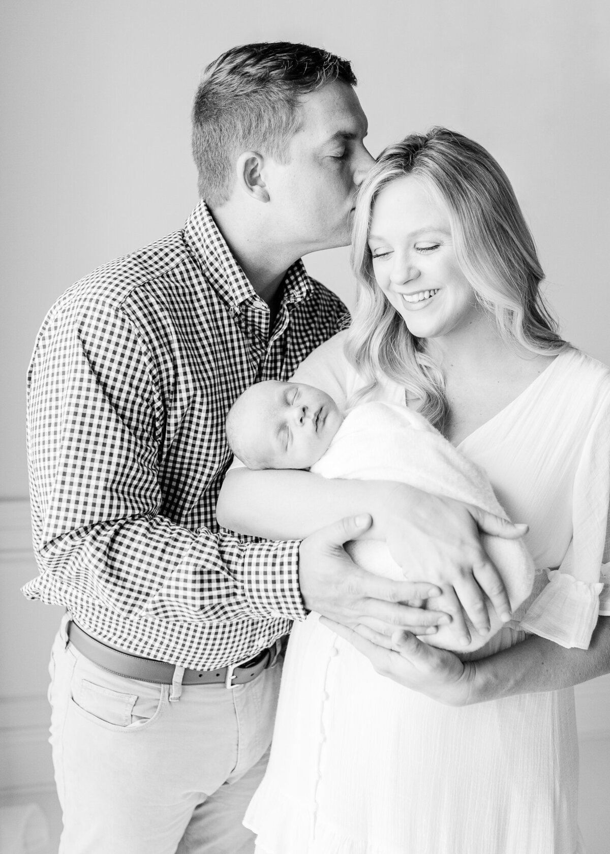 Black and white photo of mom and dad with swaddled baby boy smile together in white studio for studio newborn session. Image captured by Brandon, MS studio newborn photographer.