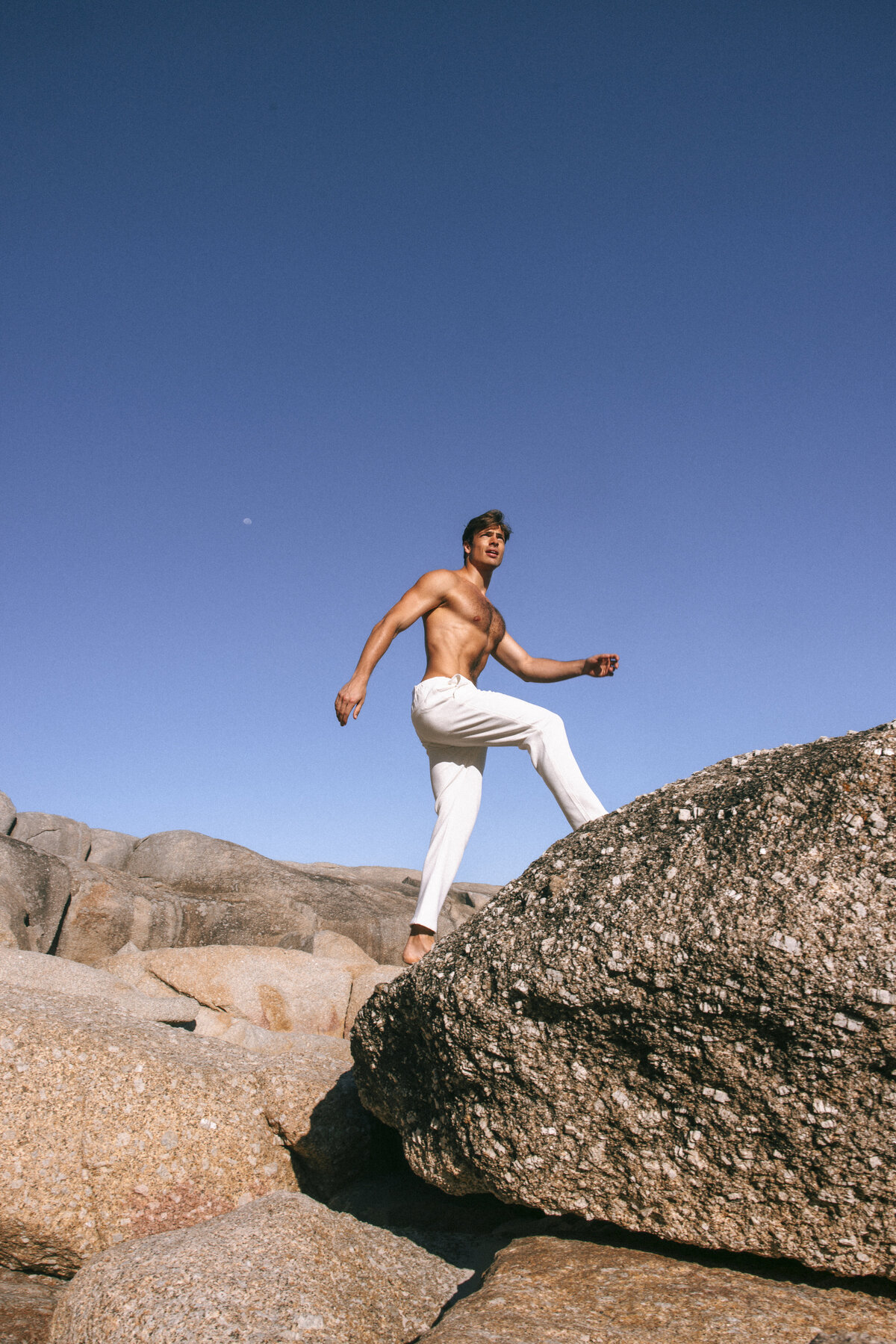 Shirtless male model stepping onto a rock formation in Cape Town, wearing white trousers under a clear blue sky, with the moon faintly visible in the background.