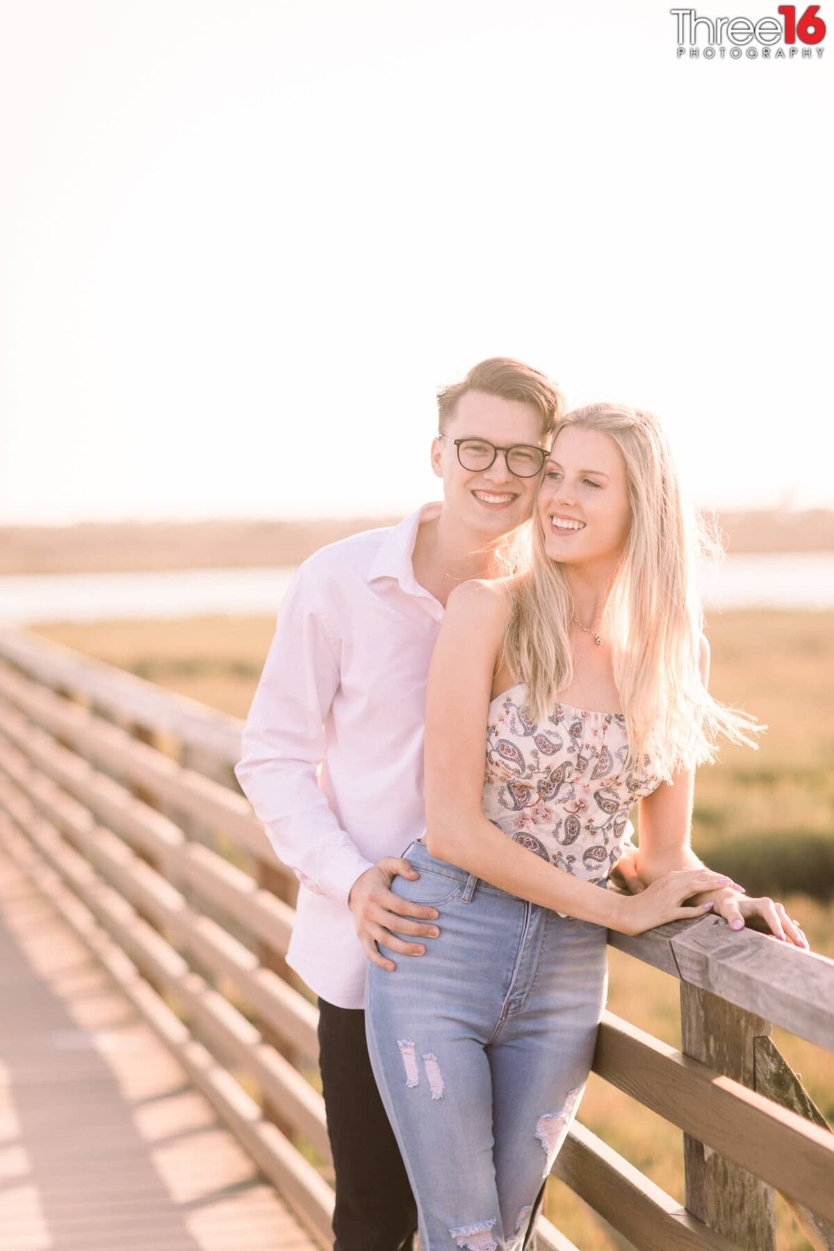 Engaged couple pose together leaning up against a ranch style fence