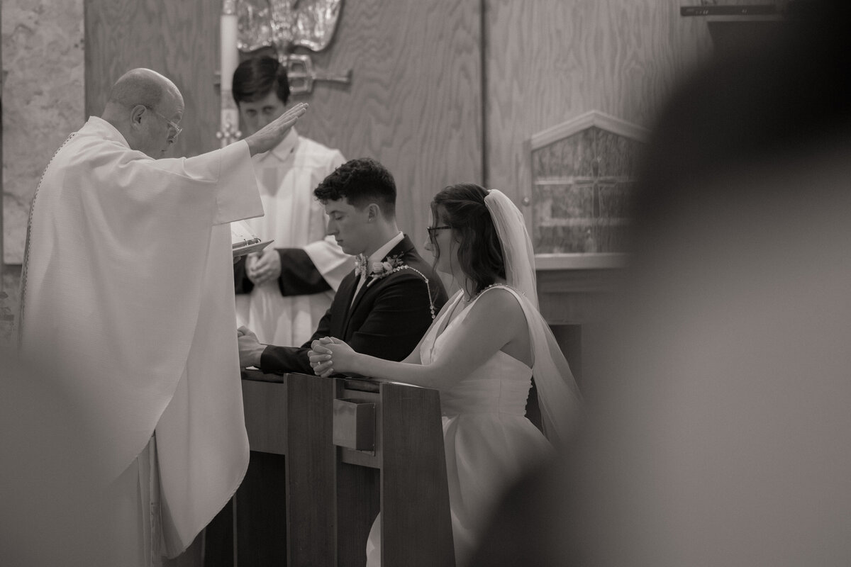 A priest blesses a bride and groom during a Catholic wedding ceremony. The couple kneels in prayer at the church altar, capturing the solemn and spiritual moment of their marriage vows.