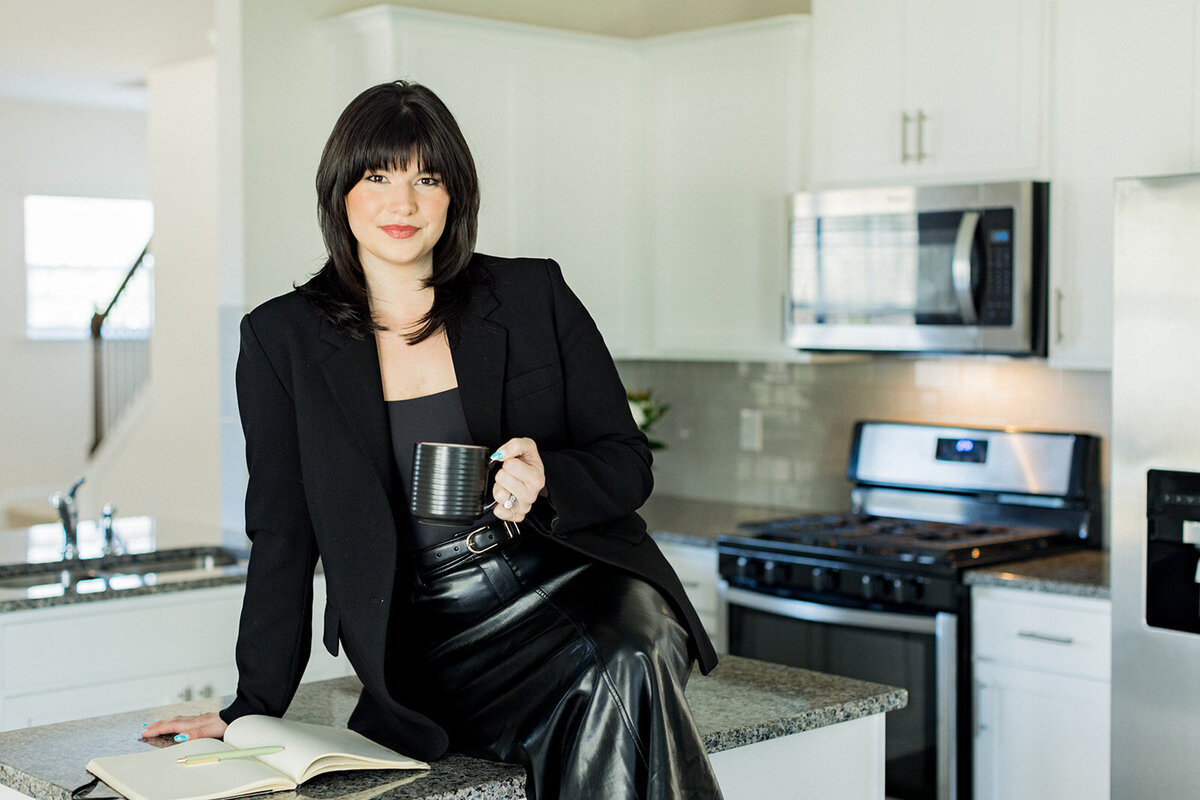 Columbia, SC business branding photo of Olivia Cooley from Olivia Cooley Real Estate, dressed in a black blazer and leather skirt, sitting on a kitchen counter with a coffee cup and open notebook, captured by Jessica Hunt Photography.