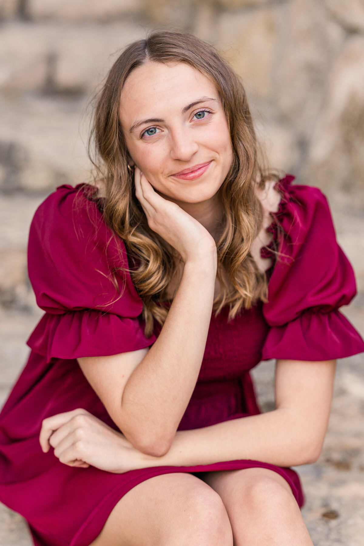 Close-up senior portrait of a young woman in a burgundy dress seated on stone steps at Adriatica Village in McKinney, Texas. She rests her face gently in her hand and smiles softly at the camera, capturing a warm and natural graduation moment.