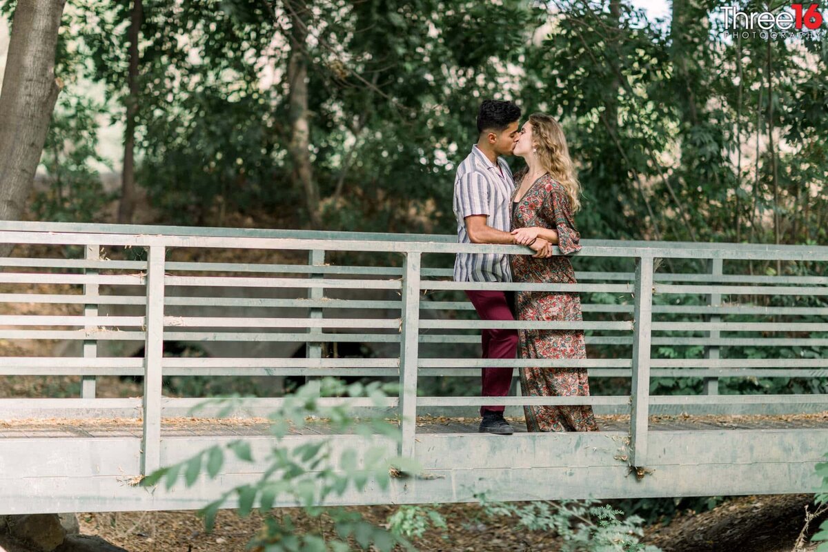 Engaged couple stand against railing on a bridge sharing a kiss with each other