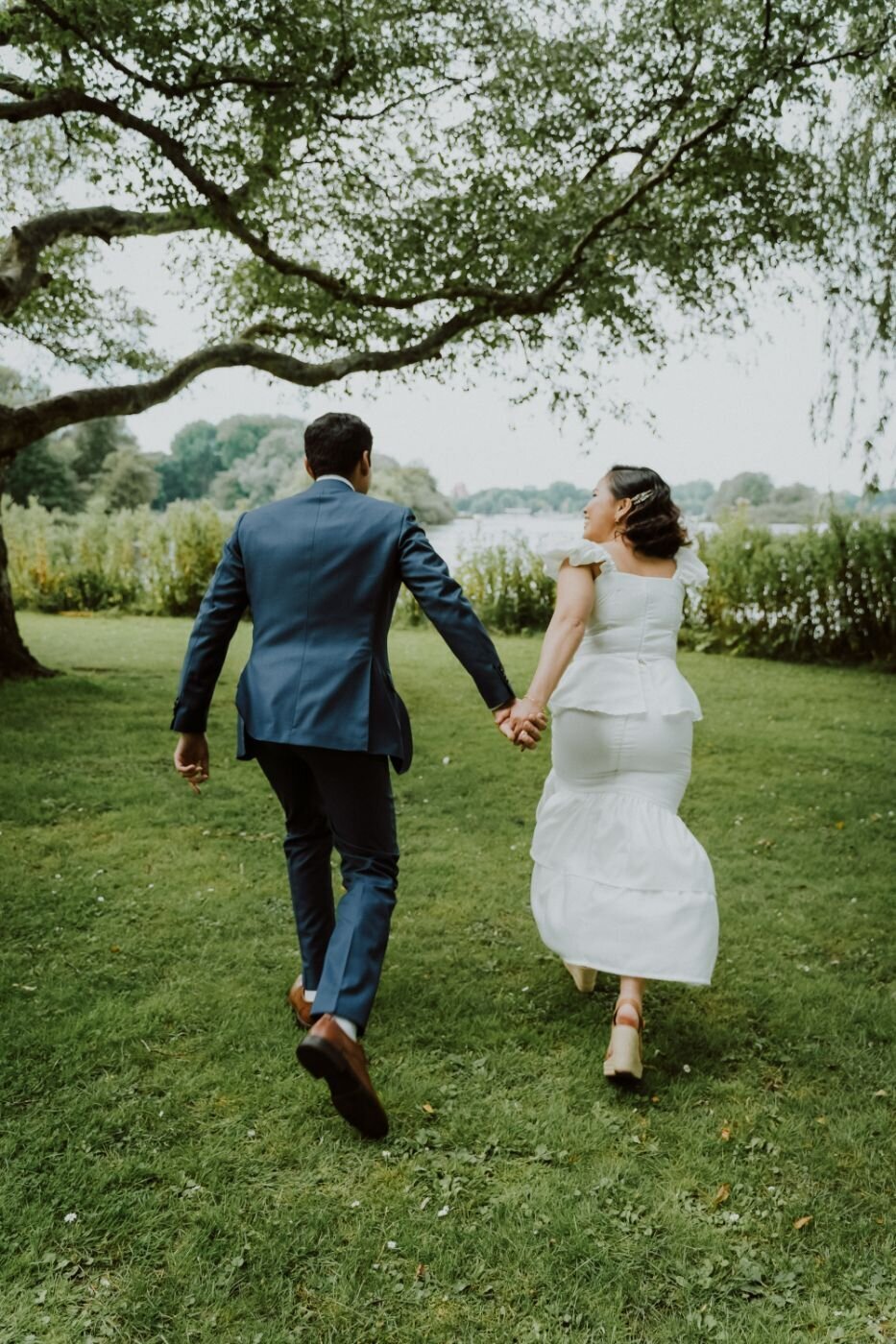 A photo of a couple from behind, running hand-in-hand through a grassy park. The man, wearing a blue suit, holds the hand of the woman, who wears a white tiered dress and heels. A large, old tree with branches hanging low is on the left, and a body of water is visible in the distance on the right, under a gray, overcast sky.