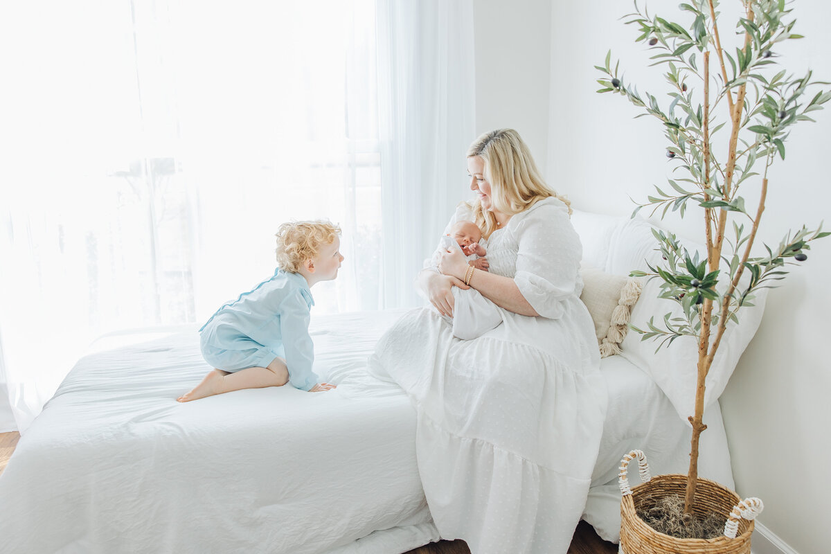 mother sitting on bed next to a window holding newborn while big brother looking on