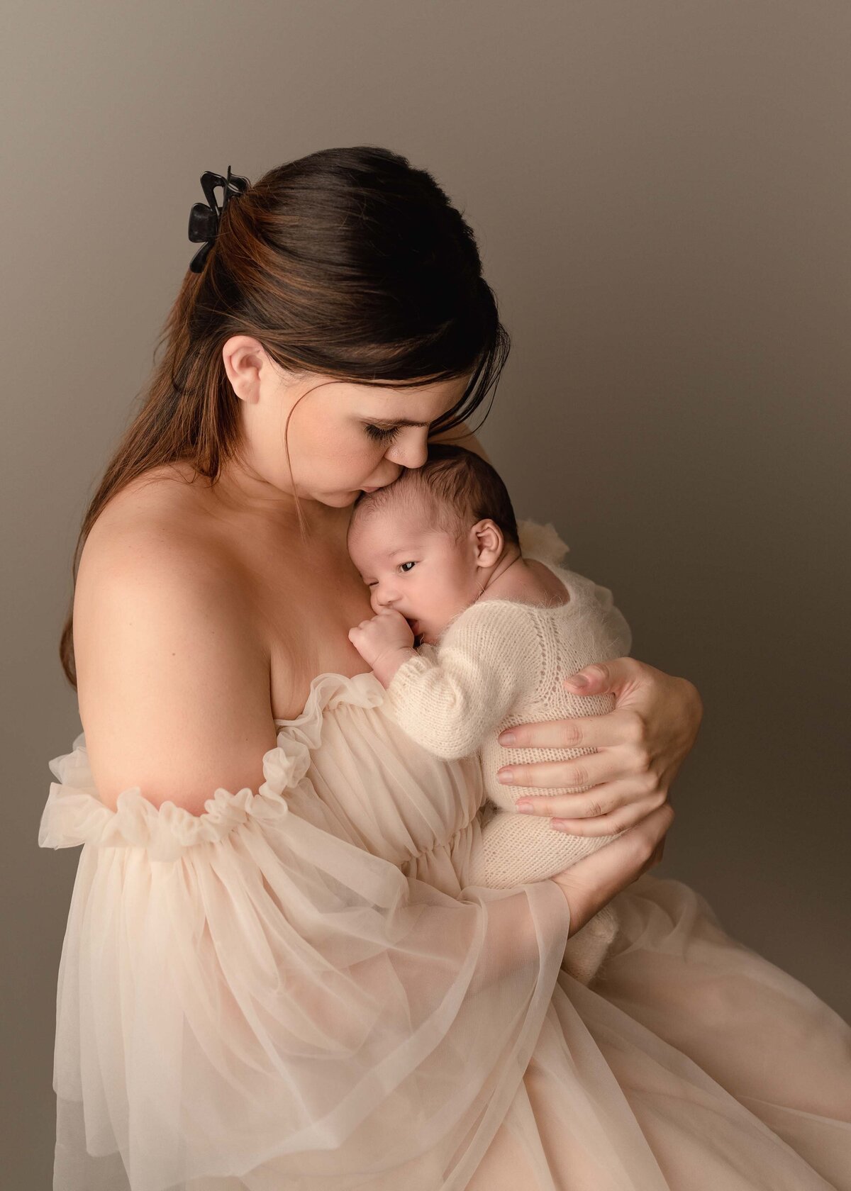 Newborn photoshoot. Mom is wearing an off-the-shoulder organza gown and holding her baby boy against her chest. Mom is giving baby a kiss atop of his head. Captured by best Murrieta newborn photographer Bonny Lynn Photography.