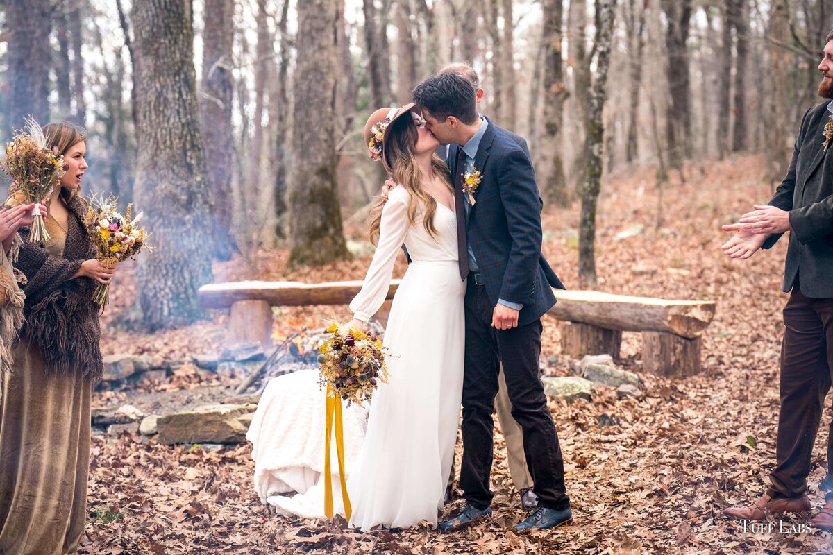 Bride and groom kiss at outdoor winter wedding ceremony and she holds her dried flowers bouquet.