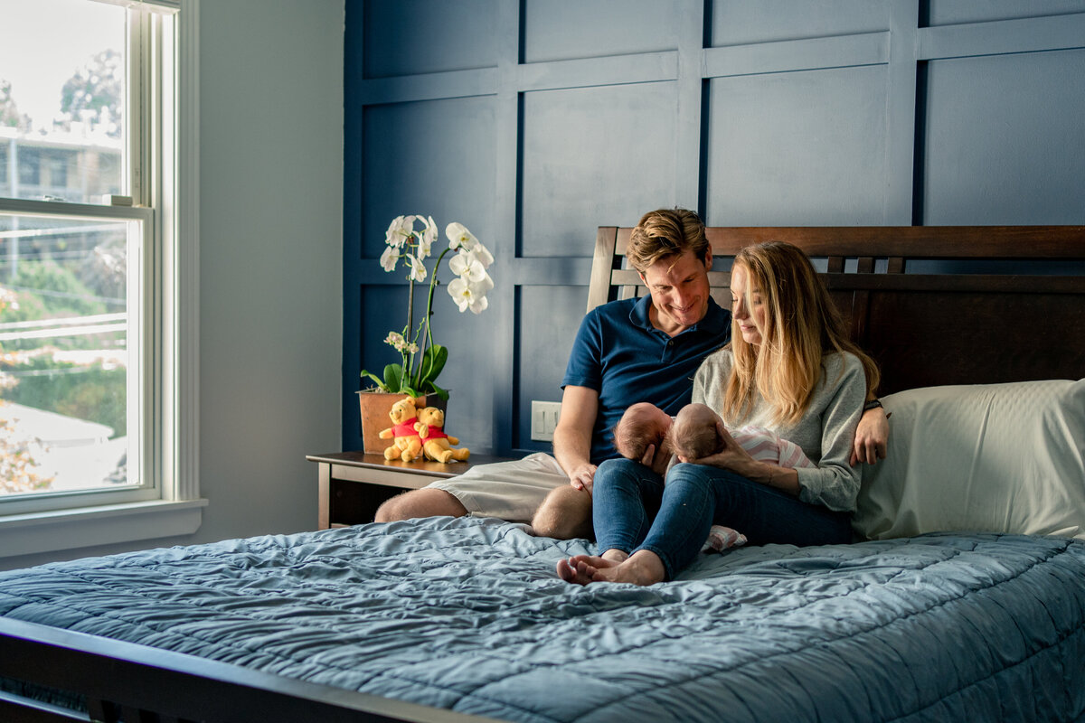 Mom and dad sitting on bed looking at little babies in their lap on their bed during in home newborn photo session