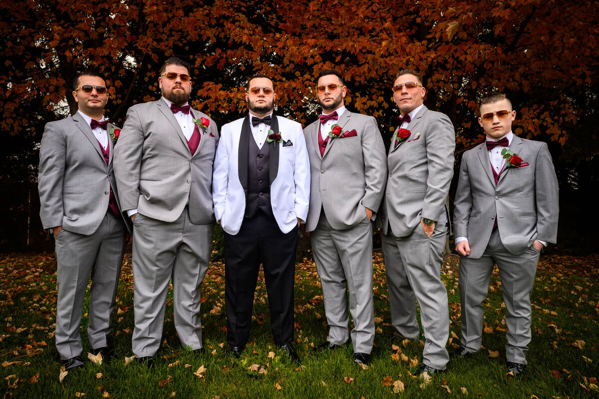 A groom in a white suit jacket poses with his groomsmen outside, who wear grey suits