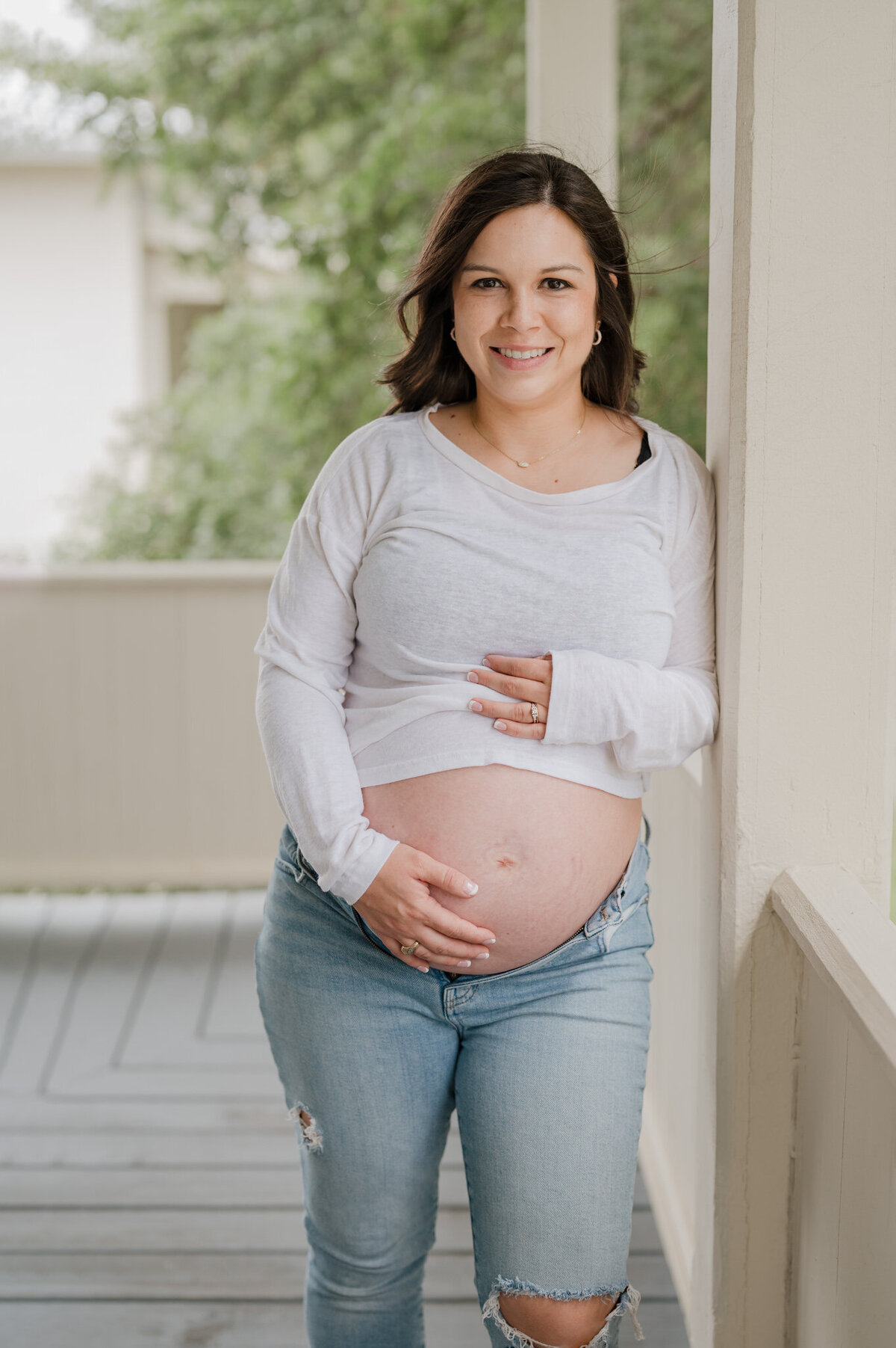 Pregnant woman in jeans and a raised white shirt holds her belly and smiles at the camera during maternity pictures.