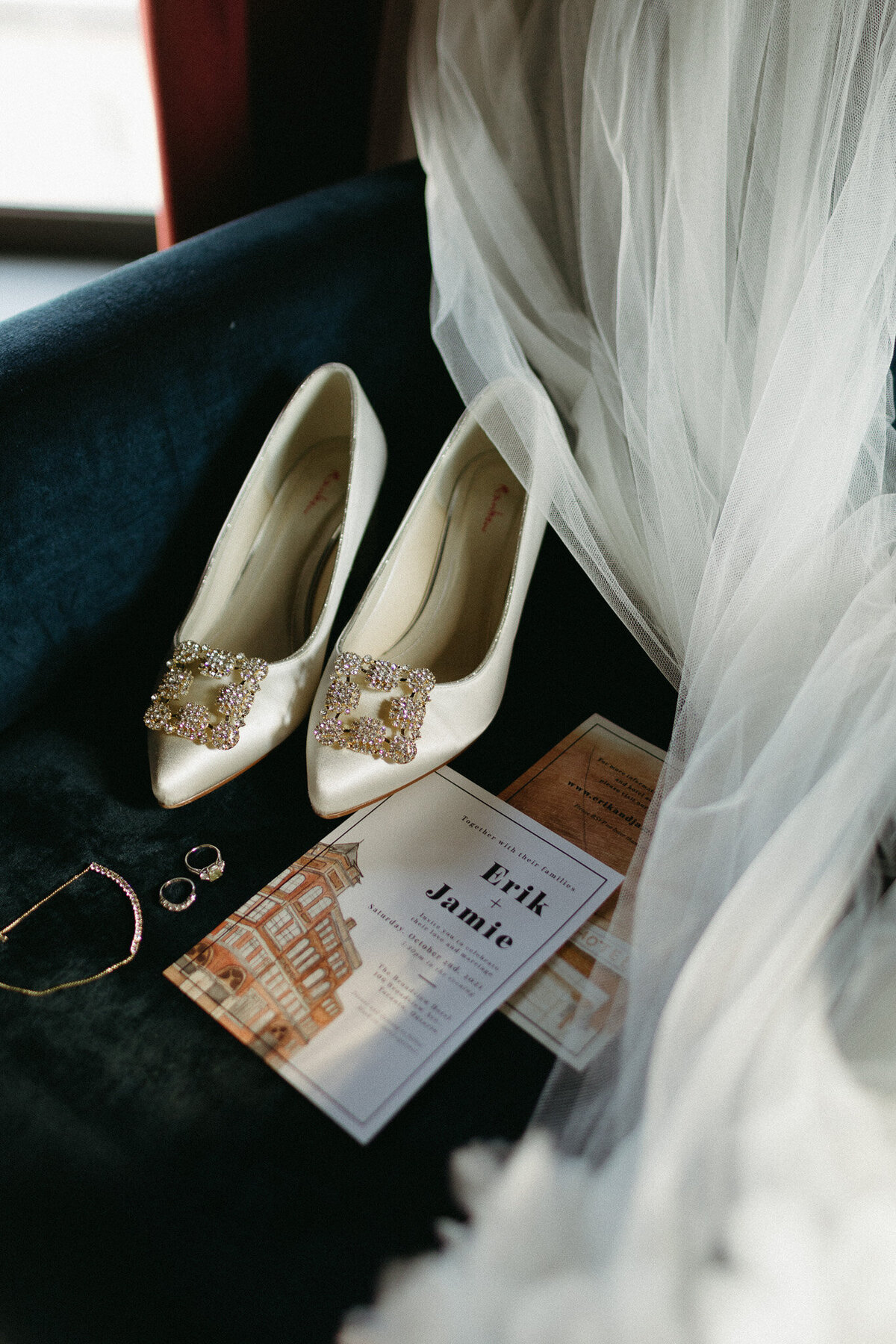 White wedding shoes with jeweled buckles and a wedding invitation are artfully arranged on dark fabric beside a sheer veil, capturing the elegance perfect for any destination wedding photographer. Nearby, two rings and a bracelet add a touch of timeless romance.