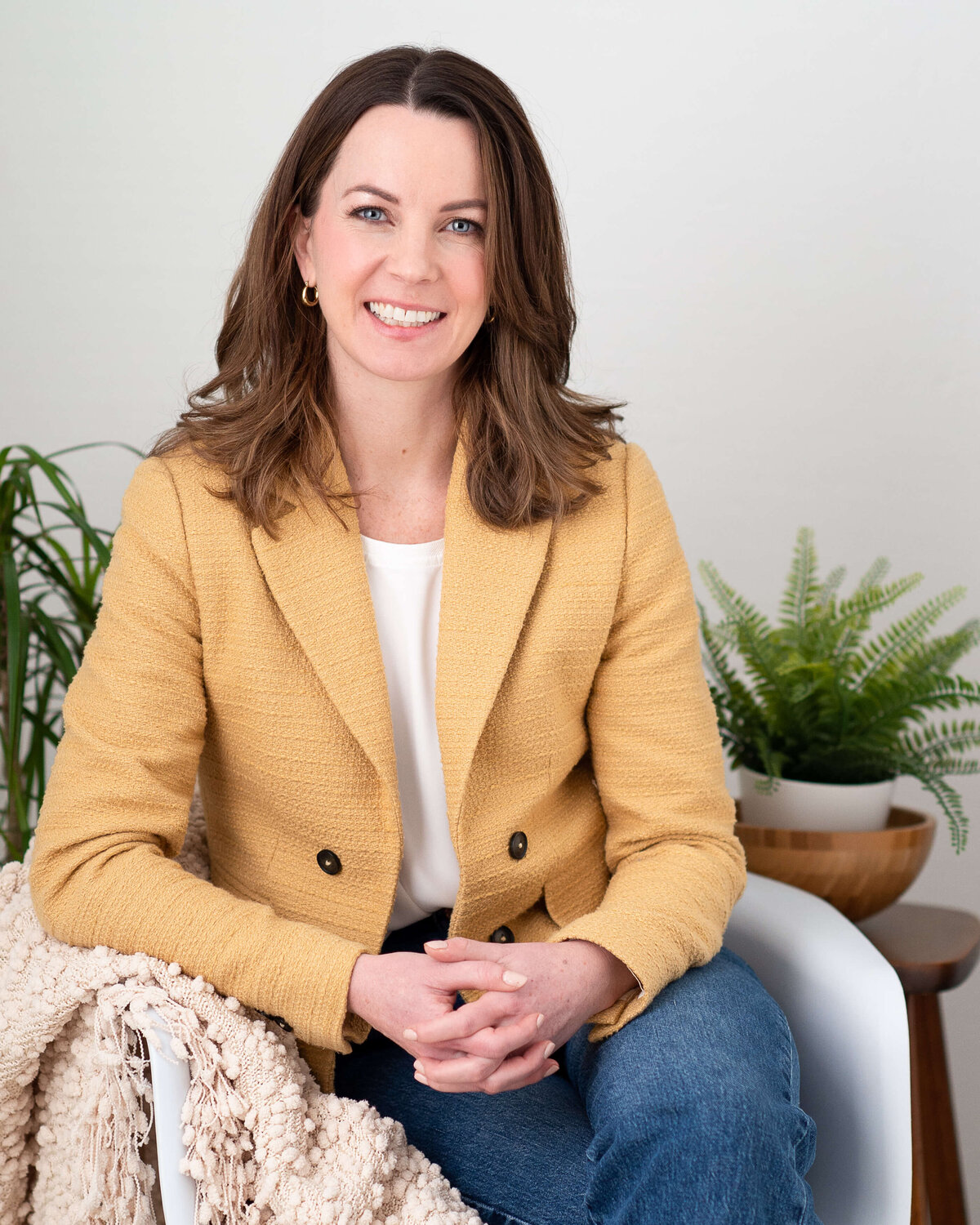 a woman in a yellow jacket sitting in a white chair with her hands clasped, smiling at the camera.  Capture by Ottawa branding photographer JEMMAN Photography
