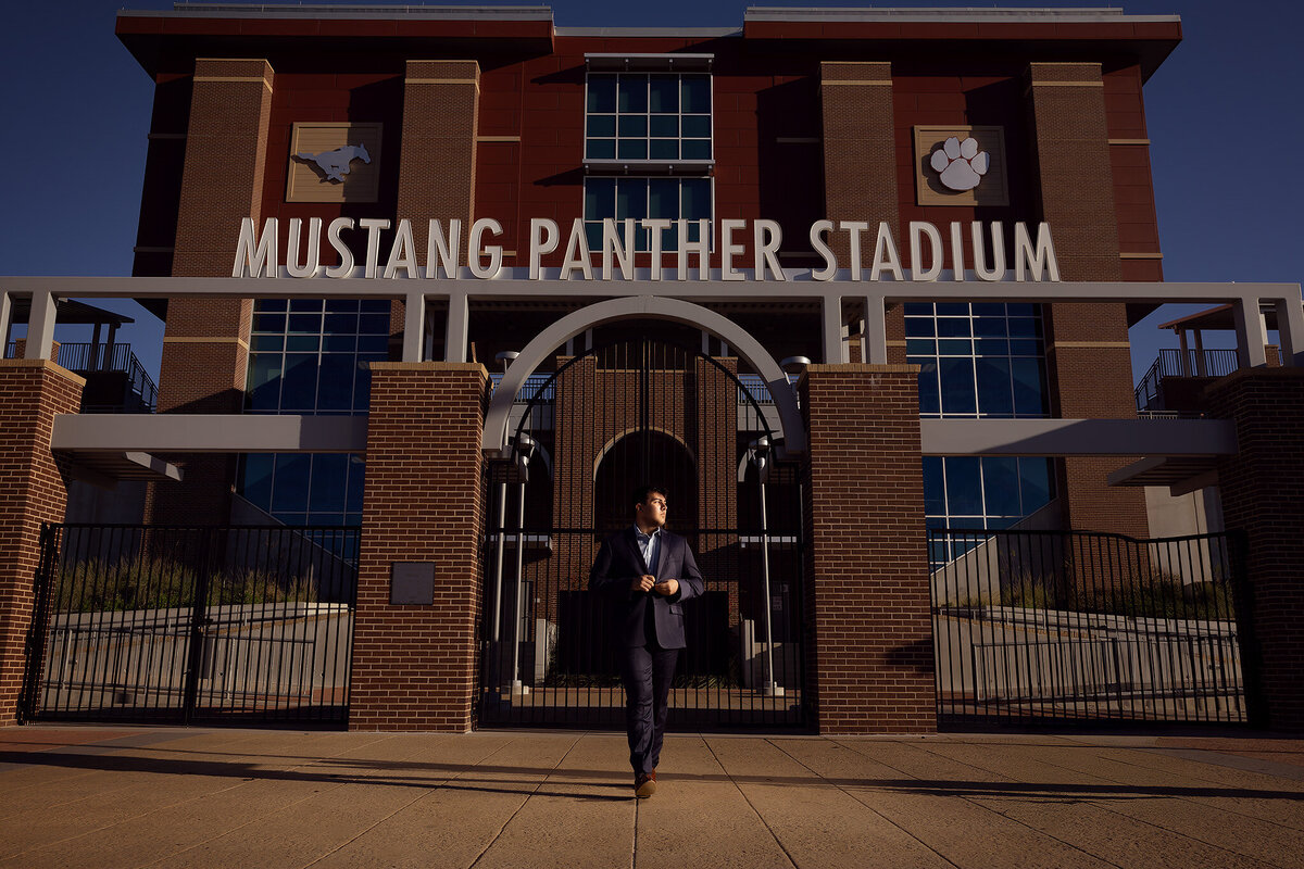 Senior wearing a suit, walking confidently in front of Mustang Panther Stadium for an inspiring Dallas senior photoshoot, blending athletic spirit with professional style. Perfect for high school senior photography in Dallas.