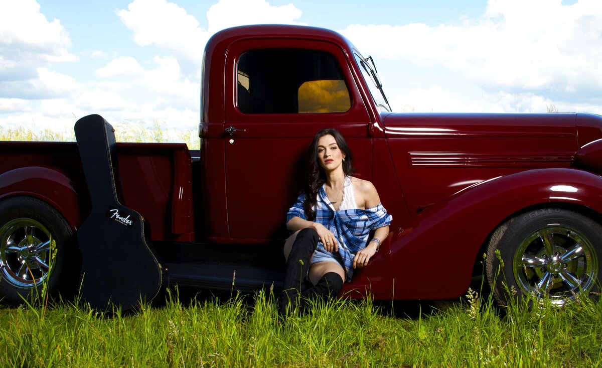 Female musician portrait Kristin Carter sitting against vintage red truck with black guitar case