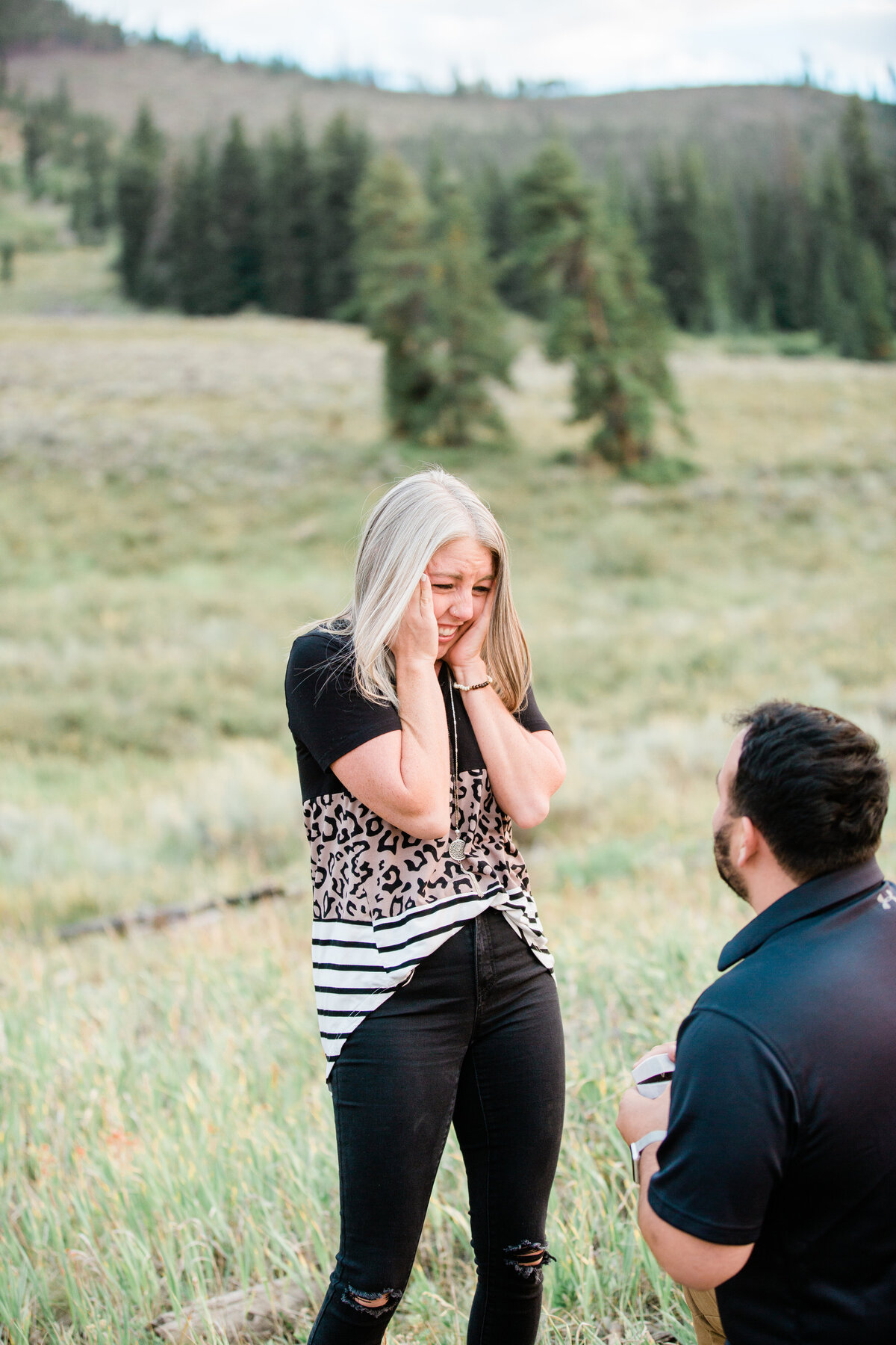 A woman has her face to her hands in excitement as her boyfriend is on one knee proposing in front of her