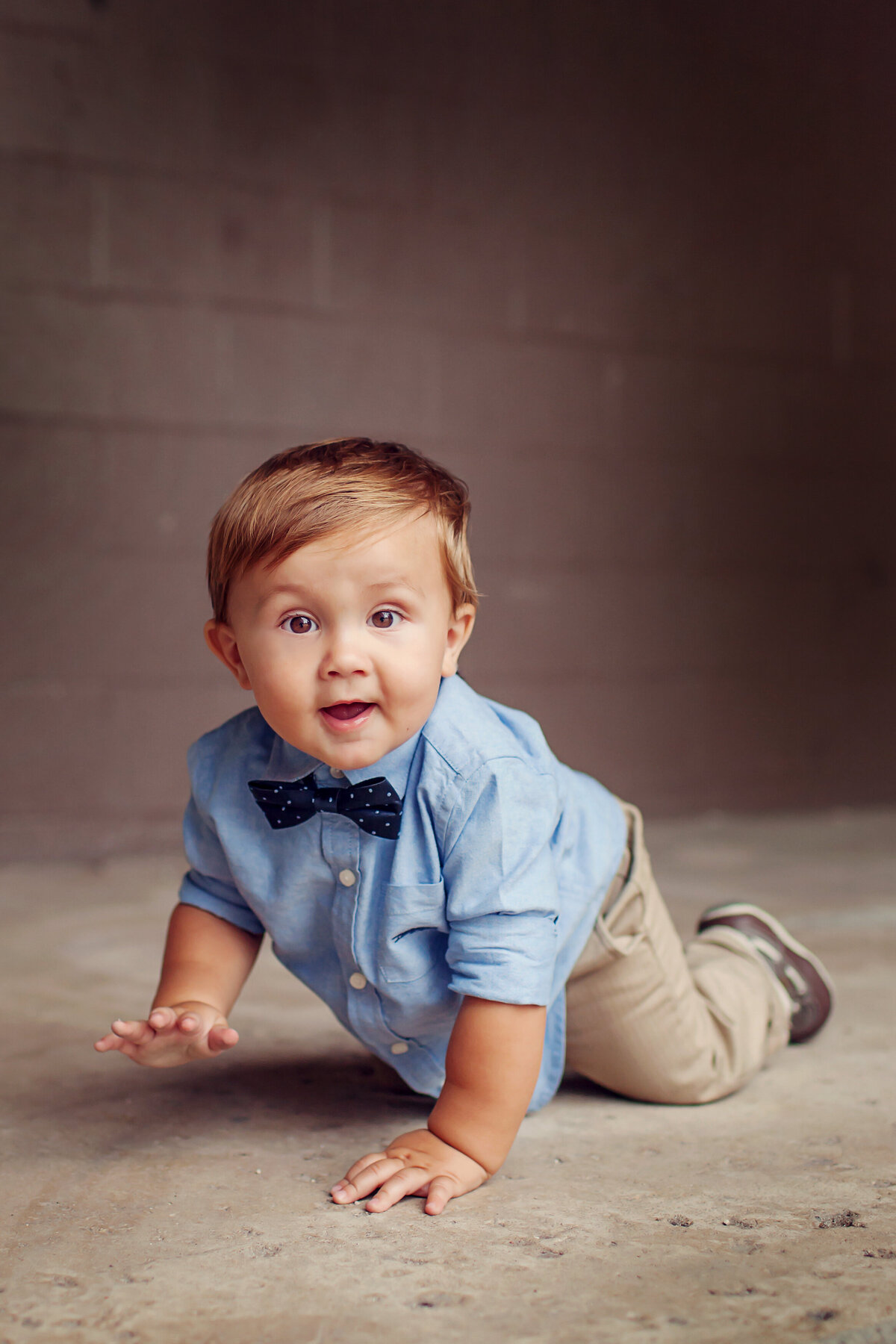 Cute little boy crawling on concrete with cubby arms and big brown eyes smiling at the camera.
