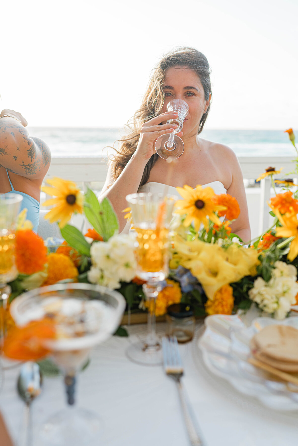 girl sipping champange with hair blowing in wind in the forefront is a white, orange, blue and green floral table runner, a champagne glass half full and an empty martini glass that housed a fruit parfait 