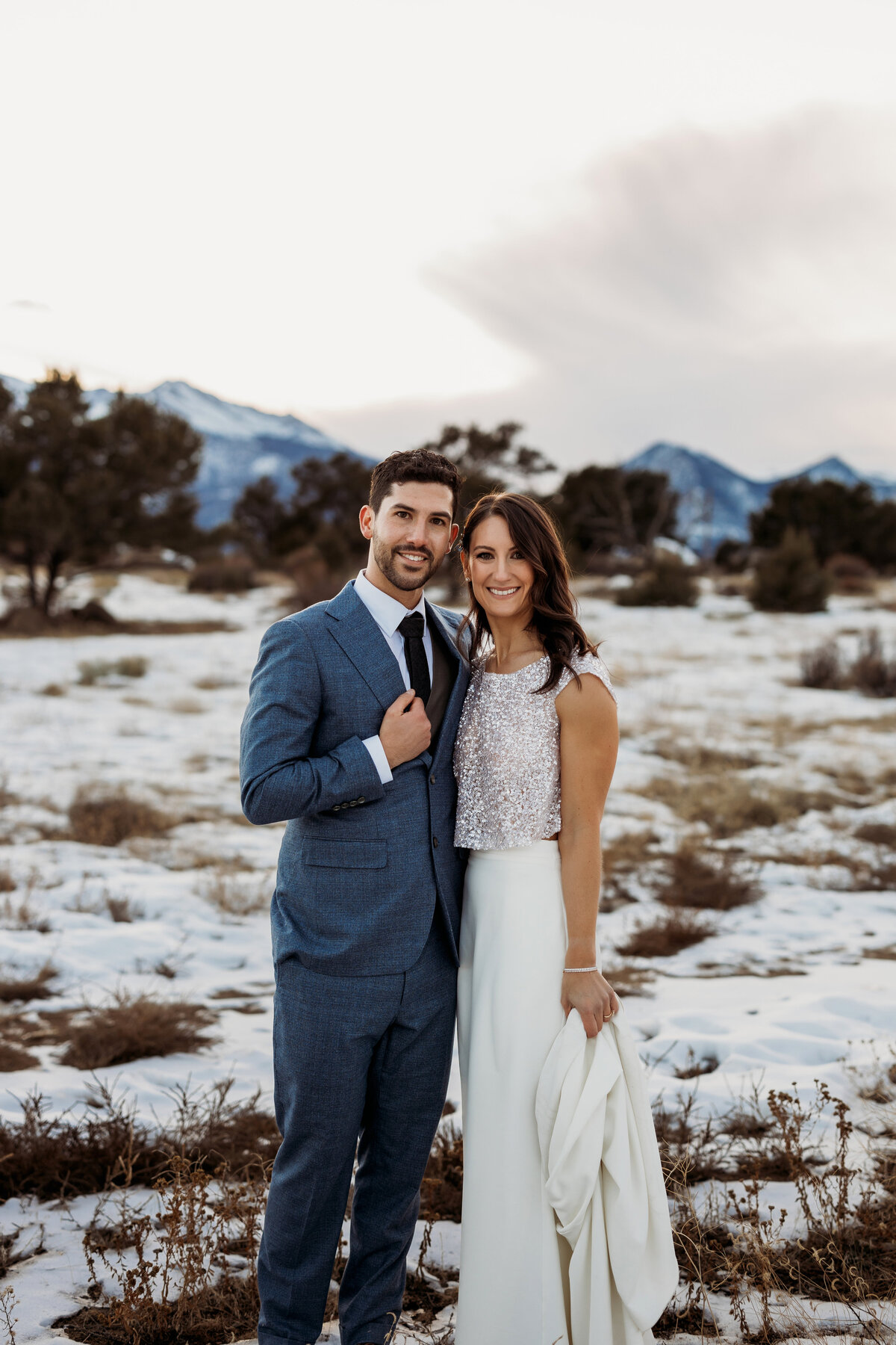 Husband and wife standing in snowy field with snow capped mountains behind them