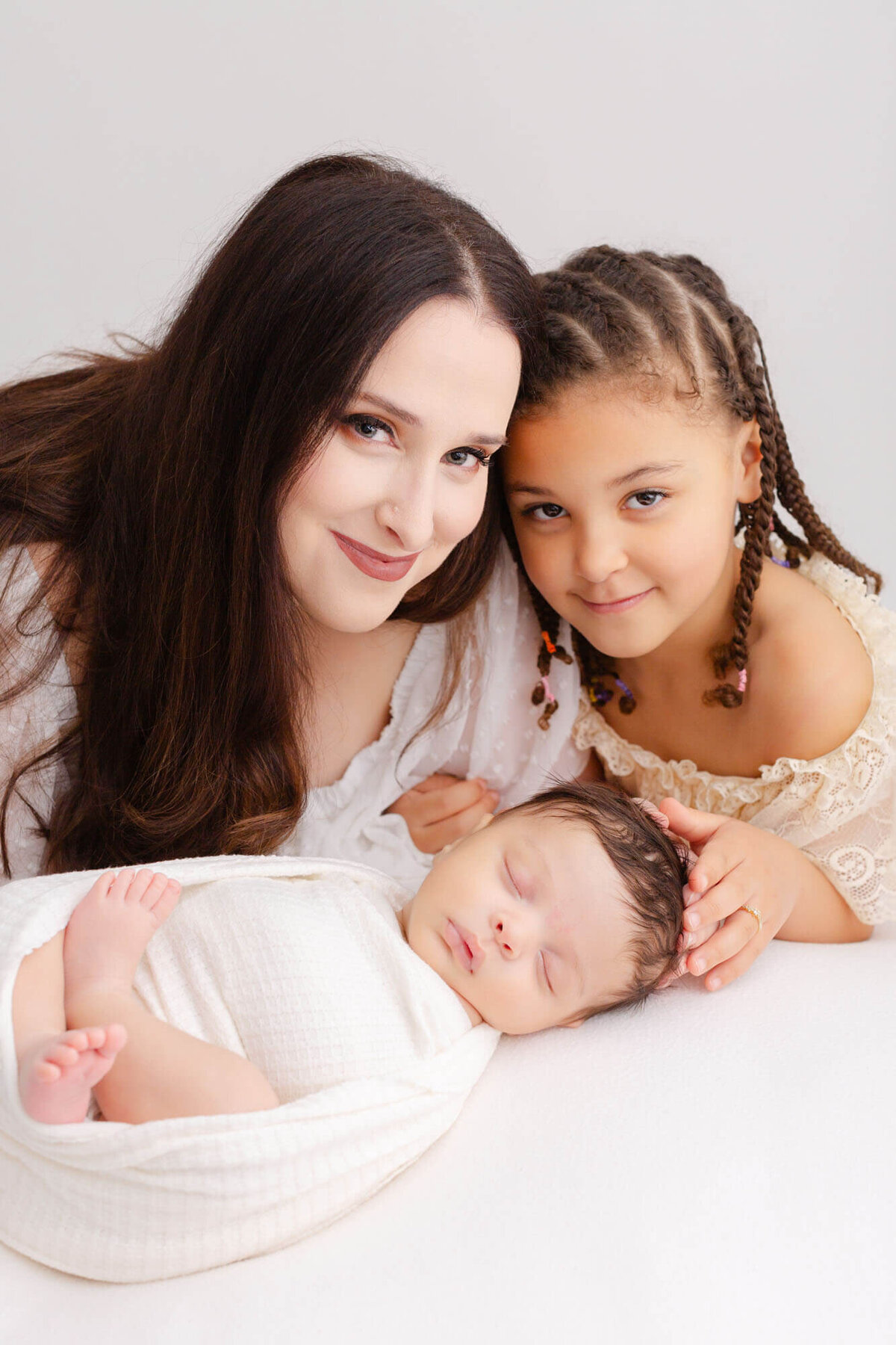Mom and toddler daughter with heads touching are looking at the camera with soft smiles on their faces while they kneel behind their newborn baby who is sleeping in front of them.