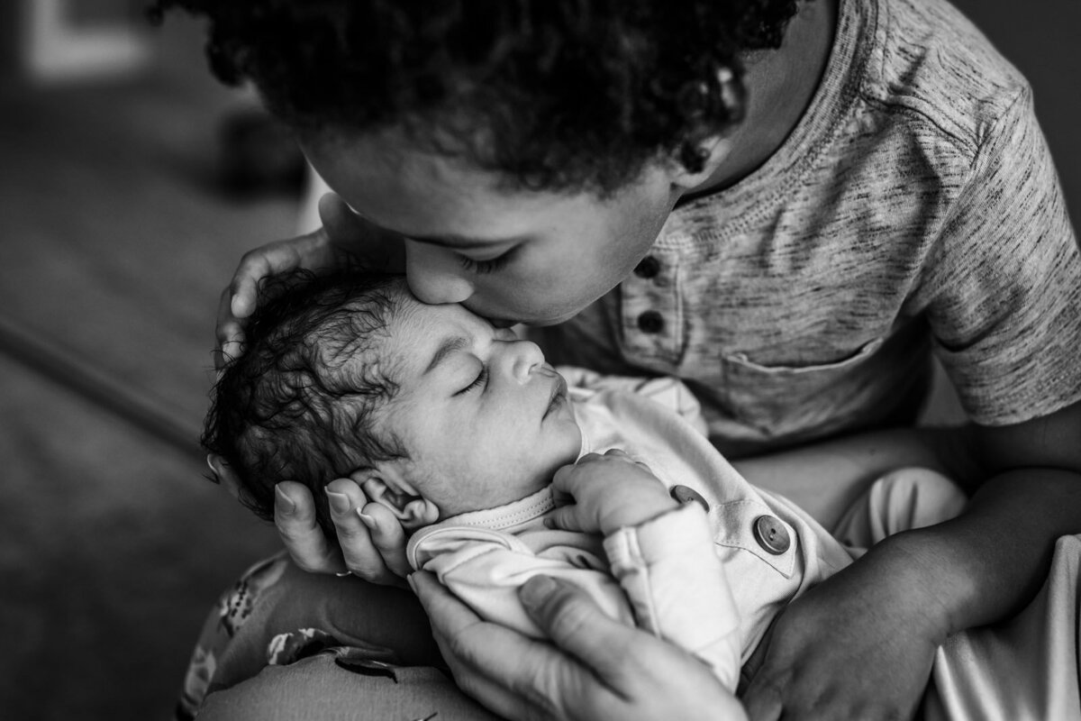 brother leaning over and kissing newborn sister held in Mom's arms