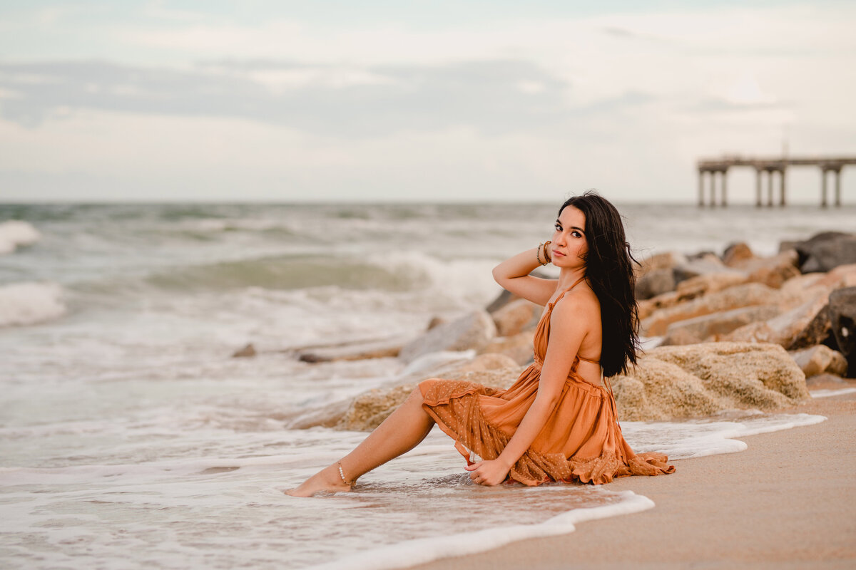 Senior photos on beach with girl sitting near the waves and rocks.