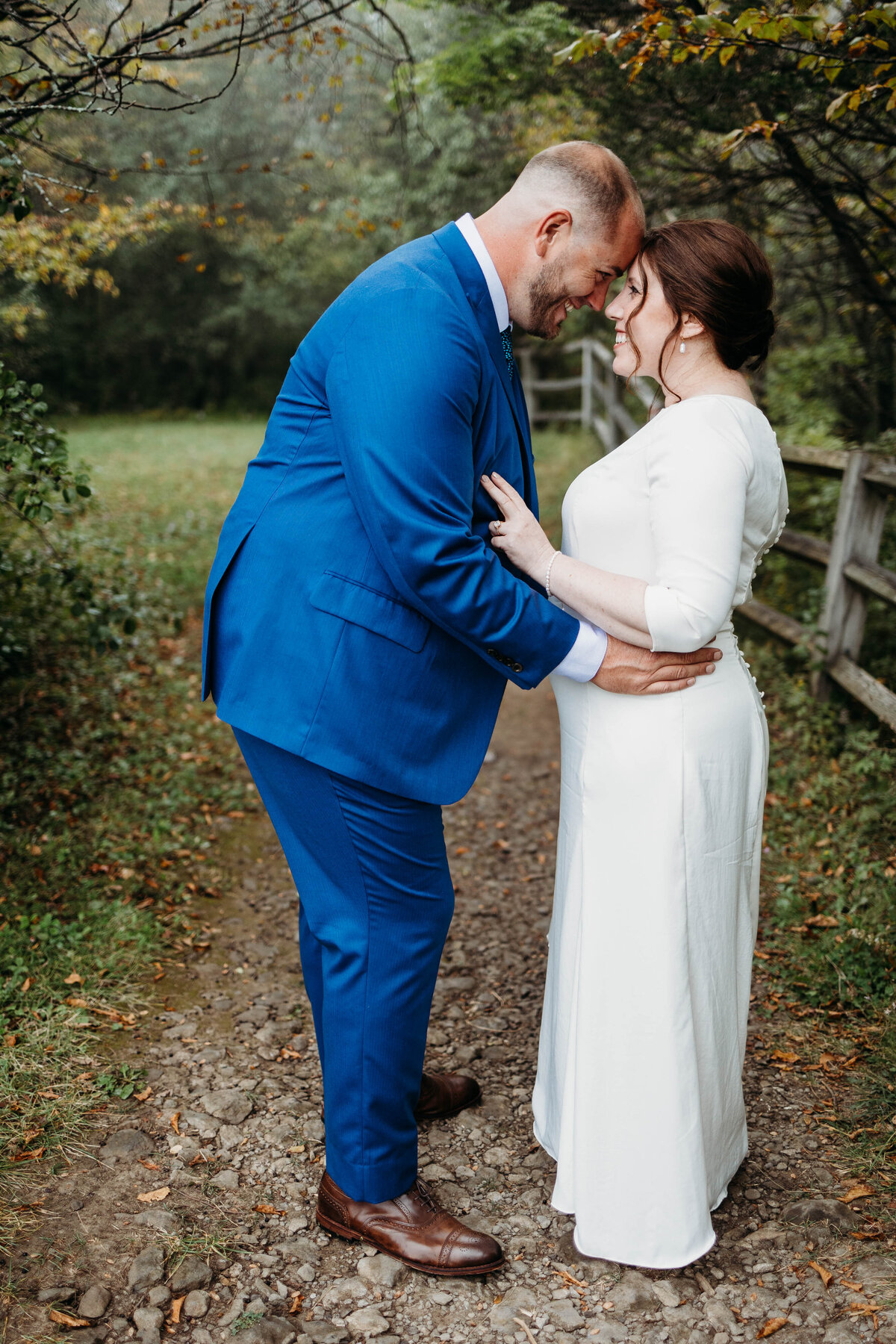 Newlyweds smile and touch foreheads on a trail by a rustic fence