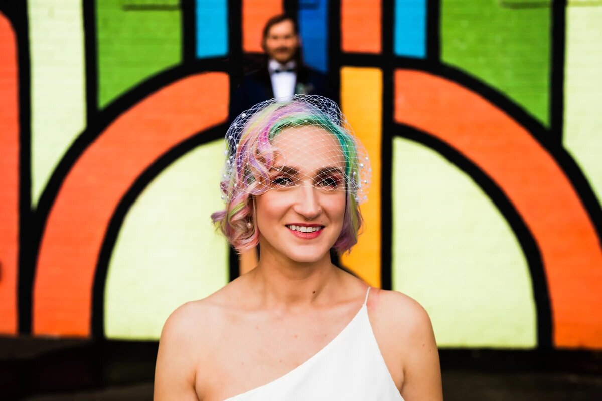 A bride with pastel-colored hair smiles brightly in front of a vibrant, colorful mural, with the groom playfully blurred in the background.