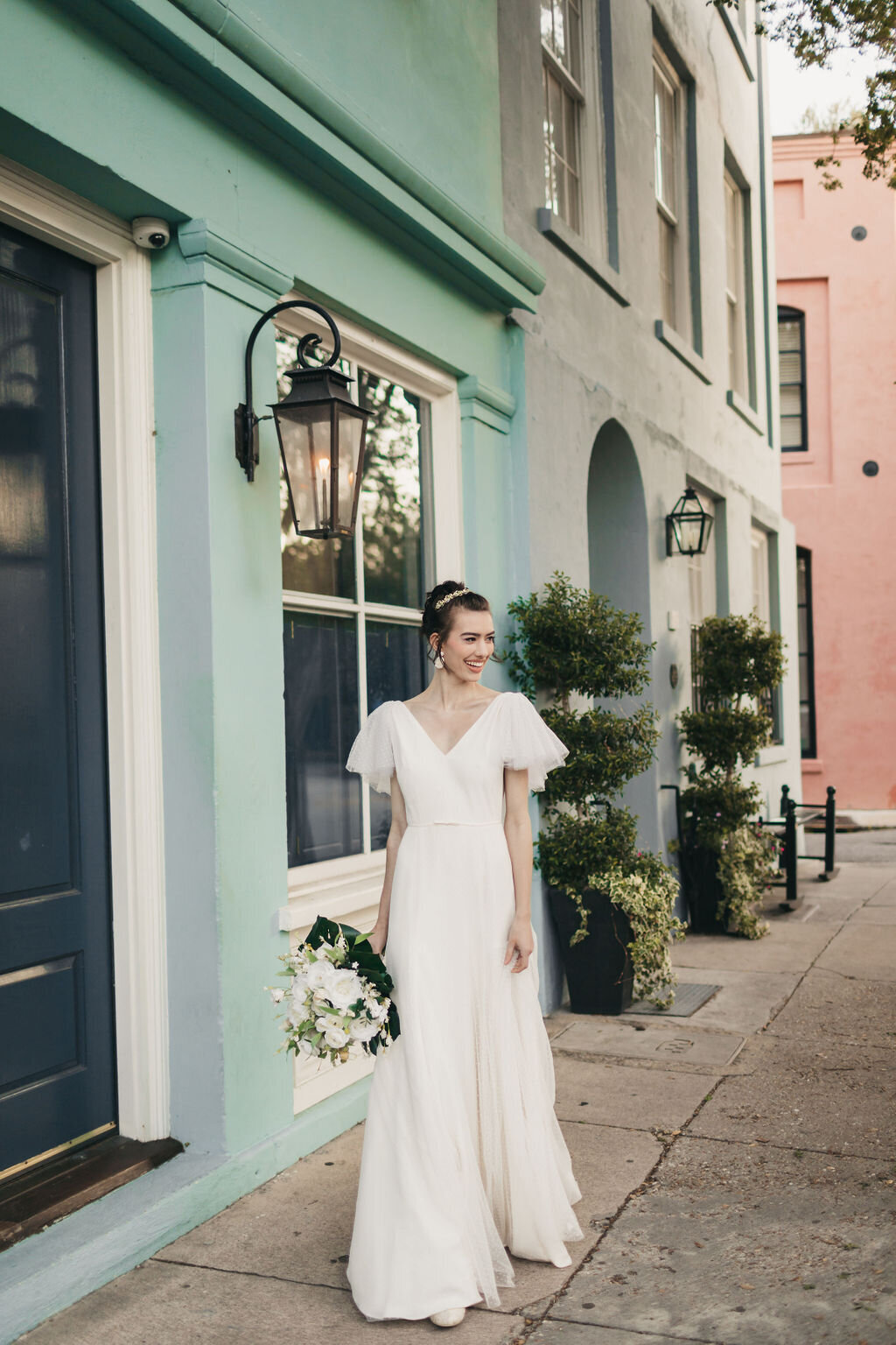 The Mai wedding dress style with its swiss dot flutter sleeves and modified a-line skirt are modeled in front of the iconic Rainbow Row on the Charleston peninsula.