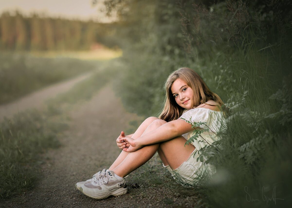 flicka sitter på en lantlig väg med högt gräs omkring sig.