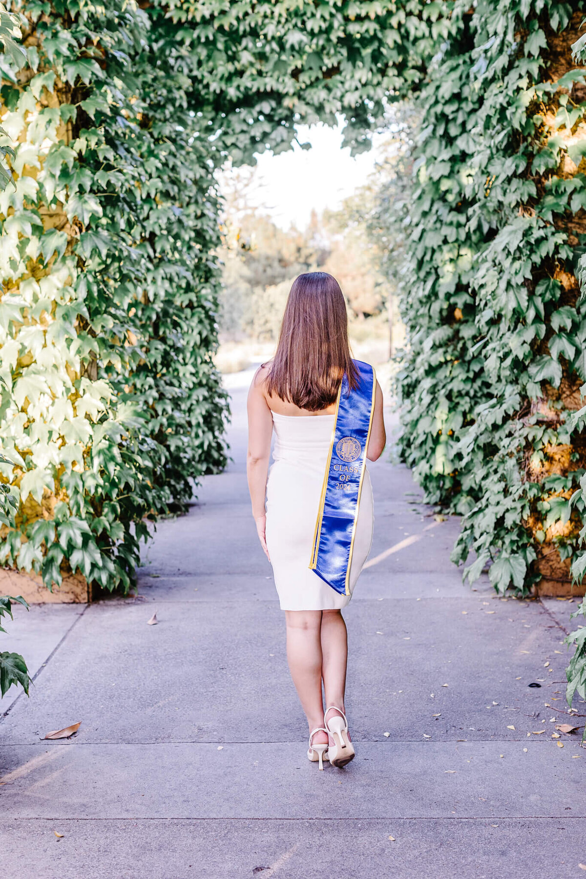 A woman in a white dress and heels, wearing a blue and gold graduation sash, walks down a pathway framed by lush green vines. She is facing away, moving toward an open area with sunlight in the background.