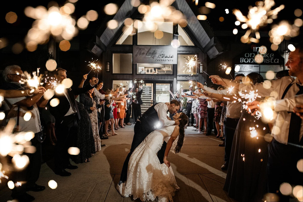 A warm and intimate wedding reception at The Lake House in Calgary, viewed from outside through large glass windows. Guests are seated at long, candlelit tables, engaged in conversation and enjoying the elegant atmosphere. The rustic charm of the venue is highlighted by stone walls, a grand fireplace adorned with greenery and candles, and antler chandeliers casting a soft glow. The dim lighting and reflections in the glass add depth to the scene, creating a cozy and romantic ambiance for the celebration.