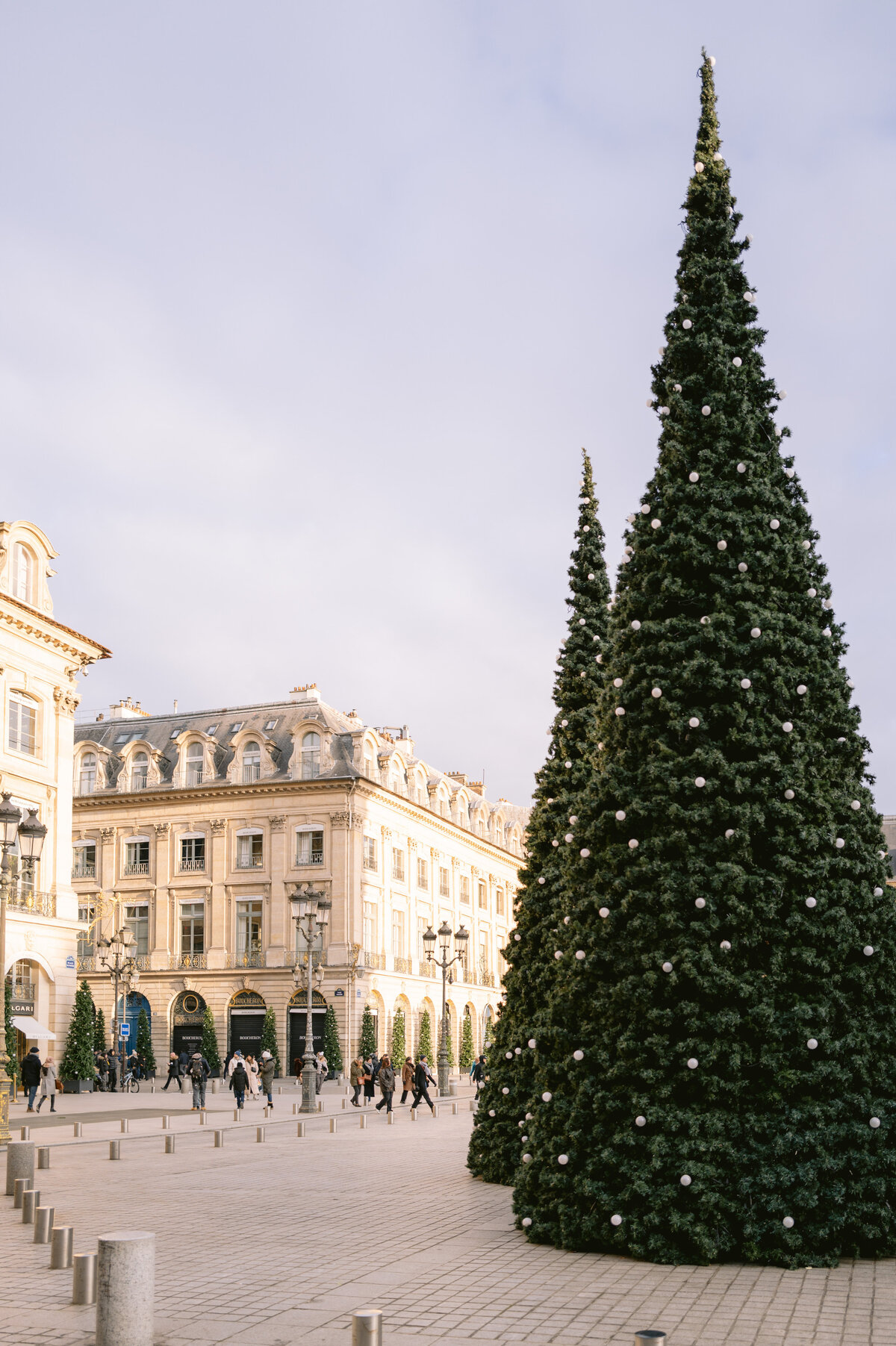 The captivating photo, taken by photographer Matteo Coltro, features a festive scene at a city square in Paris, France. The location appears to be at Shangri La Hotel & Ritz Paris Hotel, adding a sense of luxury and sophistication to the bustling holiday atmosphere. The square is adorned with two grand Christmas trees, glistening with lights and ornaments, creating a magical ambiance. Surrounding the area are elegant buildings boasting classic architectural designs, enhancing the charm of the scene. Despite the partly cloudy sky overhead, people can be seen strolling around, immersed in the joyous holiday spirit. Zen Film Works captured the beauty of this scene through their videography.