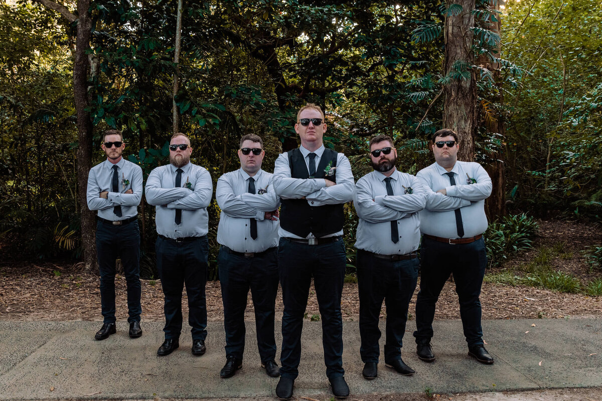 A groom and his groomsmen pose together in front of lush green trees, showcasing their formal attire and camaraderie - photo by Jamie Simmons