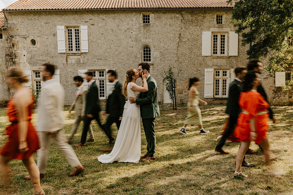 Mariés s'embrassant devant un bâtiment ancien en pierre, entourés de leurs invités en mouvement créant un effet de flou lors d'une séance photo de mariage en Vendée.