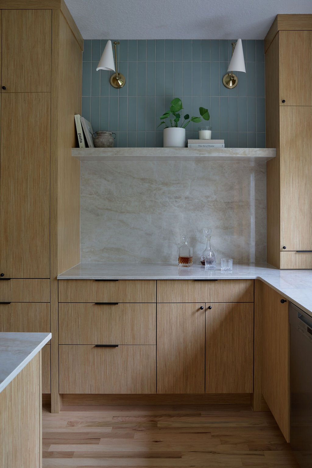 Kitchen with white oak cabinets, and slim simple black hardware. Taj mahal quartzite countertops that run up the backsplash to a quartz shelf with cookbooks and plants on it. Green subway tile backsplash above the shelf to the ceiling. Two sconces above the shelf that are modern, white and gold.